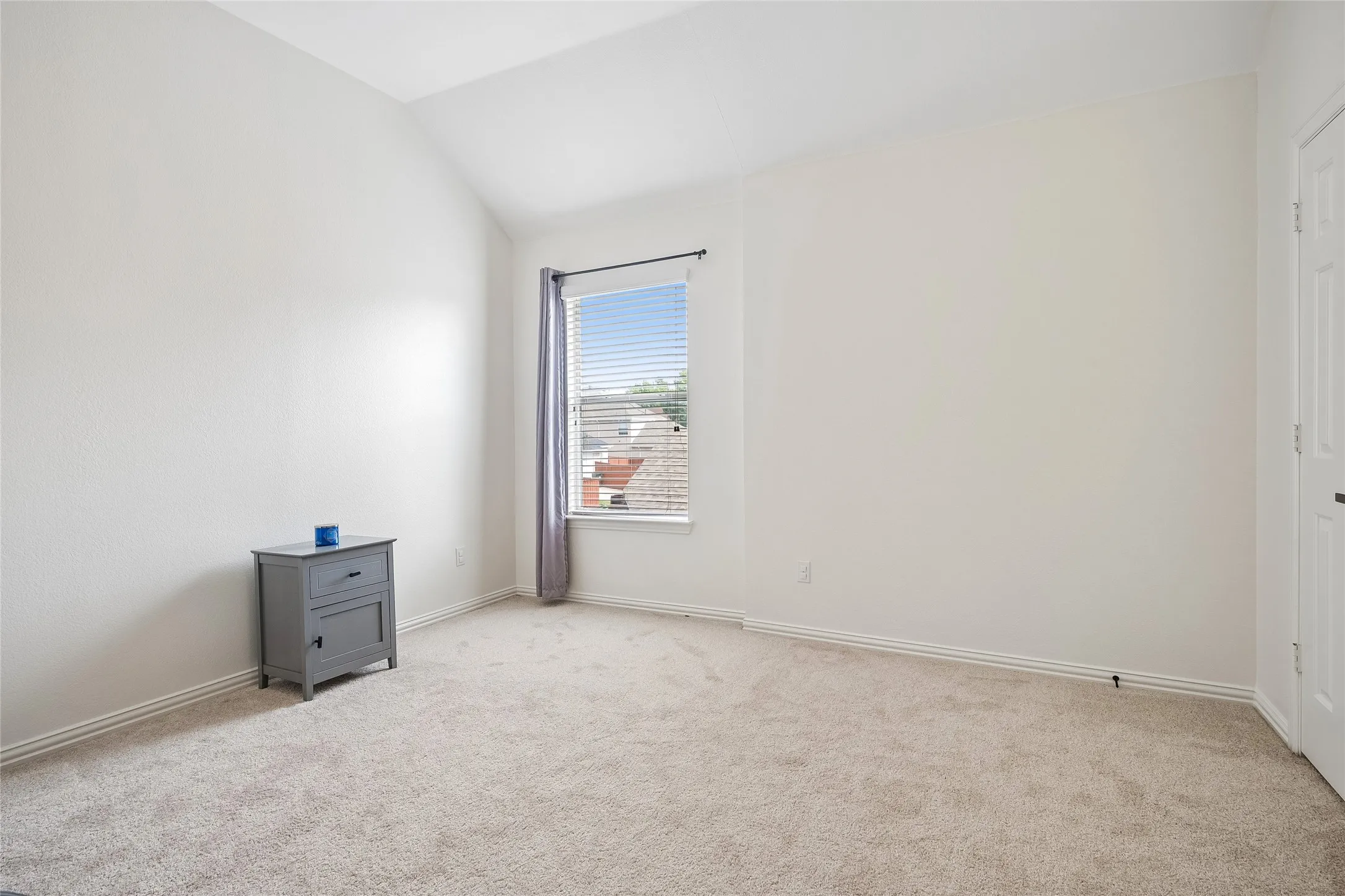 Vaulted ceiling with neutral paint, carpeted flooring, window with blinds, and white paneled door