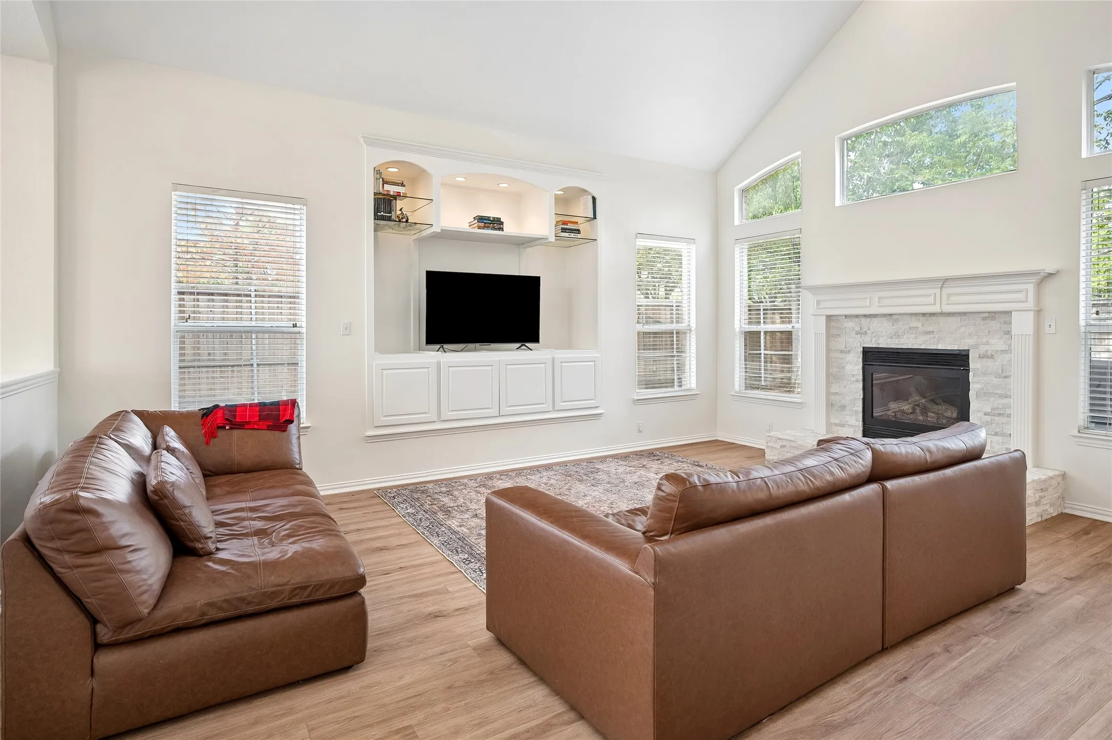 Vaulted ceiling with high windows, built-in media center with shelving, white stone-surround fireplace with a classic mantel, and wood-finish flooring