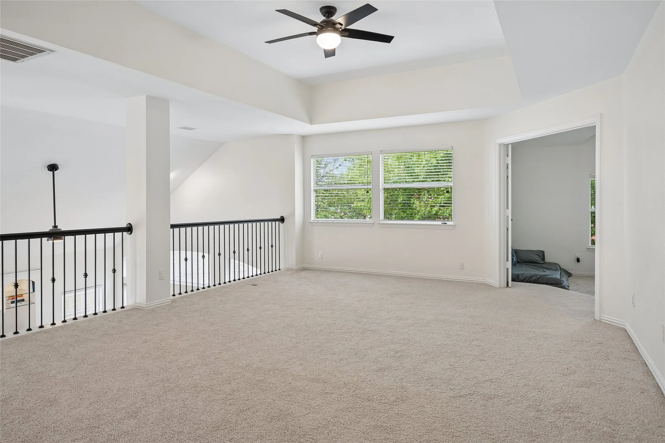 Spacious carpeted loft featuring a tray ceiling with a ceiling fan, multiple windows with blinds, and wrought iron railings overlooking the floor below
