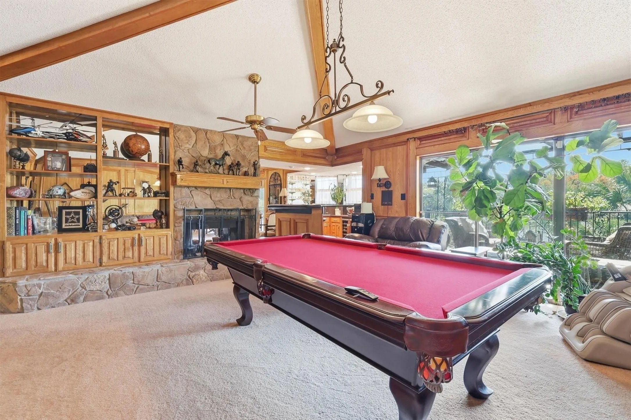 Vaulted ceiling with exposed wood beams, stone fireplace, built-in wooden shelving, wood paneling, and large windows
