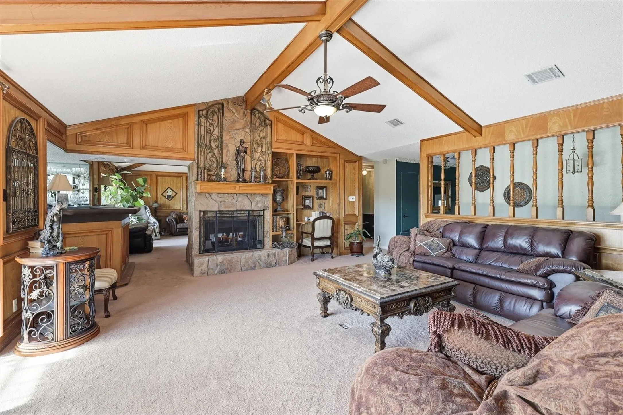 Vaulted ceiling with exposed wood beams, stone-faced fireplace with hearth, built-in shelving, wood wall paneling, and ceiling fan