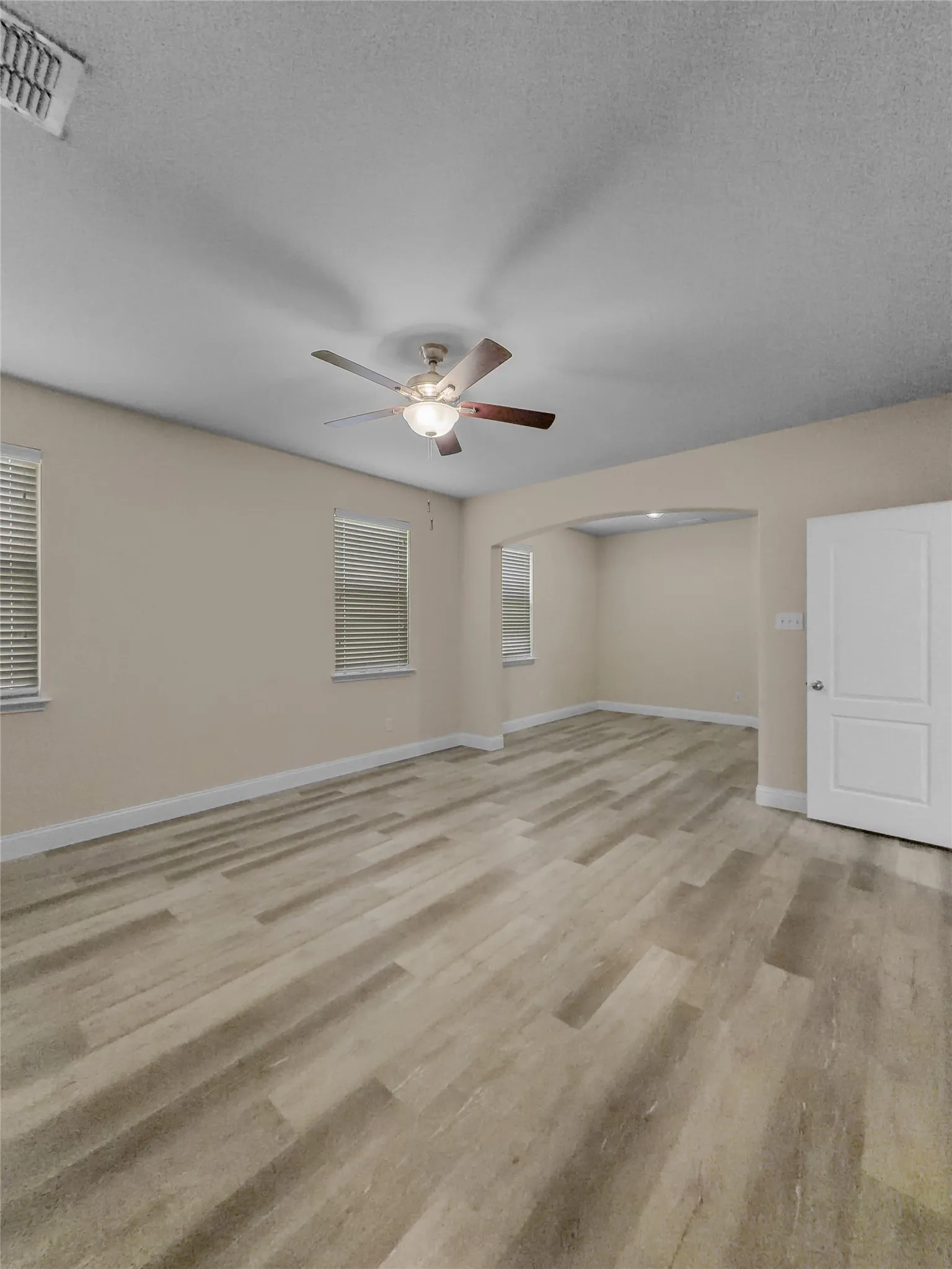Wood-finish flooring, ceiling fan with integrated lighting, multiple windows with blinds, neutral wall tones, and an arched doorway