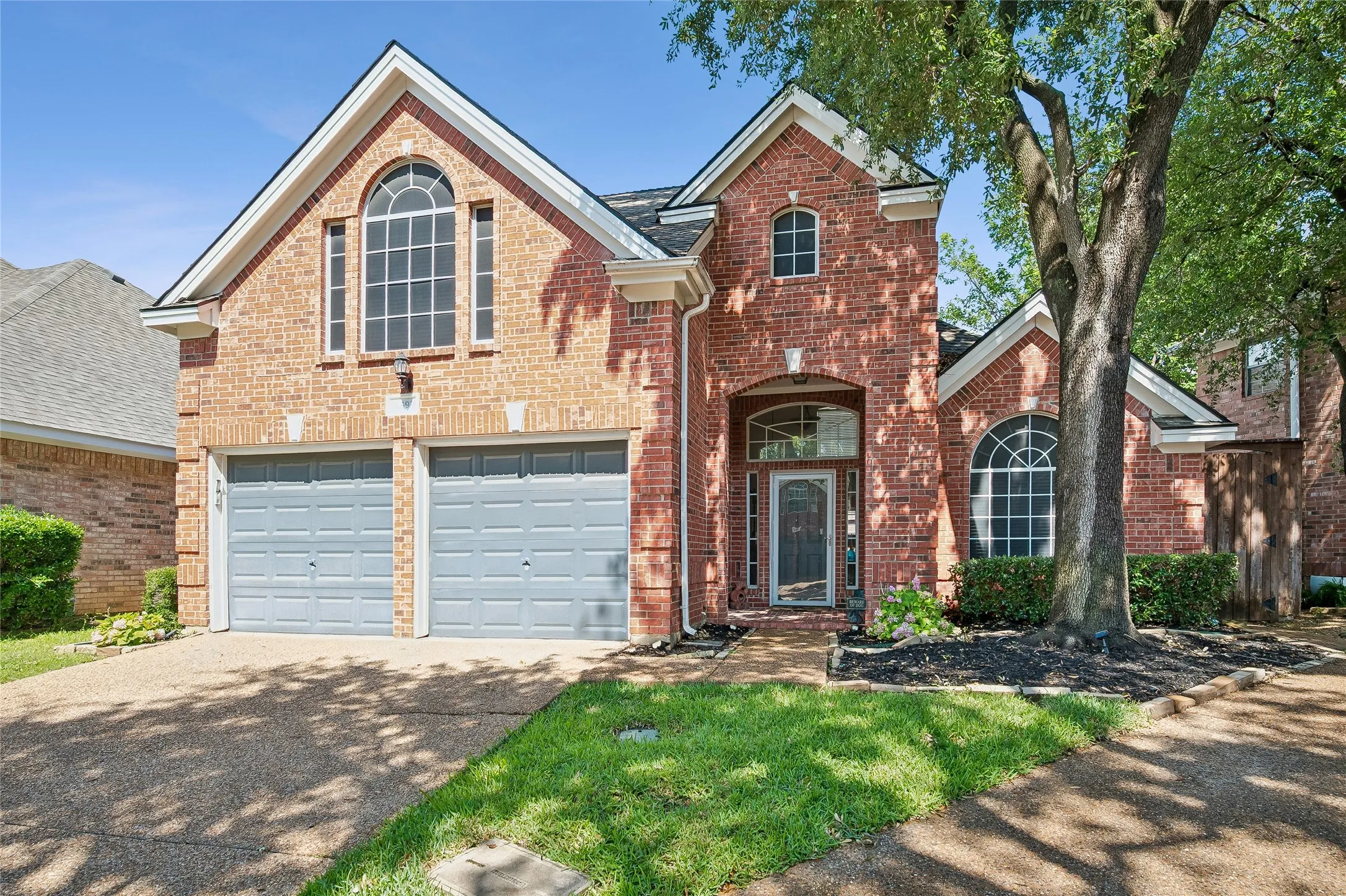 Brick exterior with white trim featuring a two-car garage, arched entryways, and a mature tree