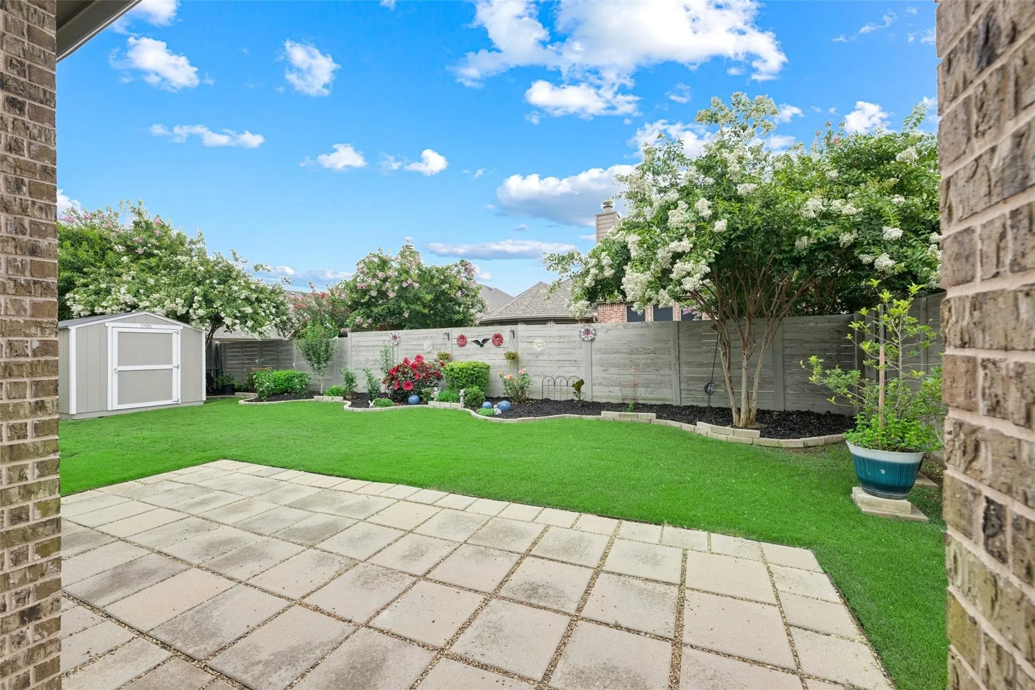 Expansive backyard featuring a rectangular paver patio, a lush green lawn, mature trees with white blossoms, a privacy fence, and a storage shed