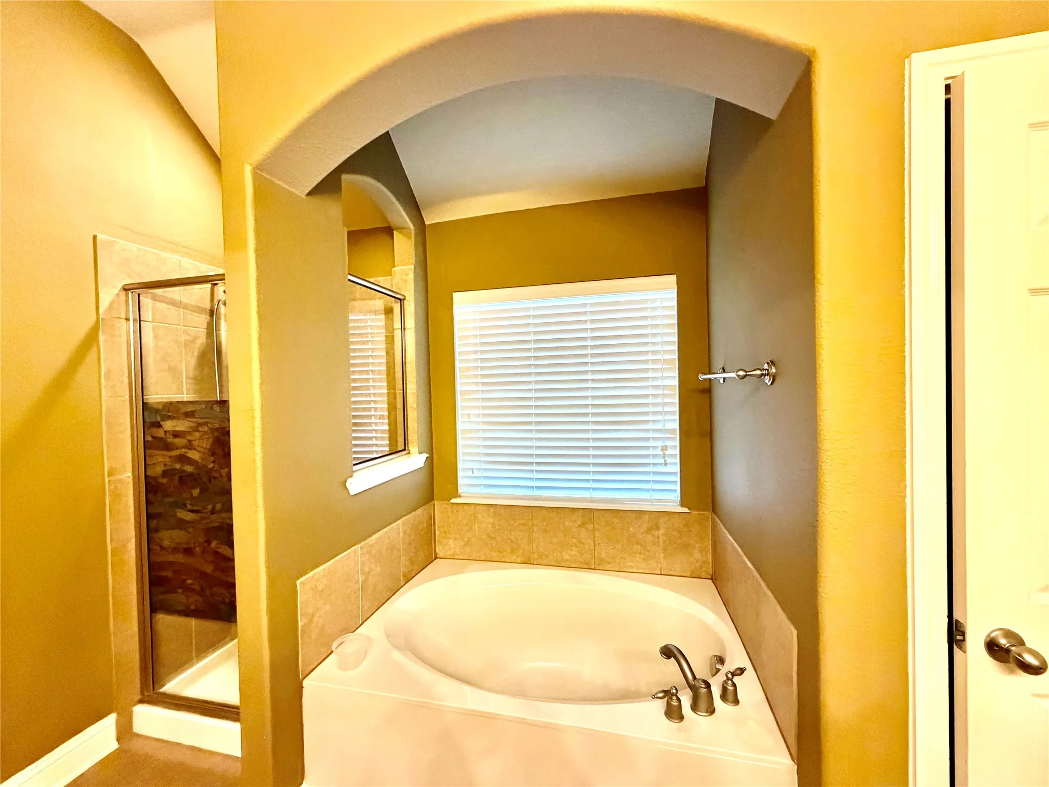Built-in corner bathtub with a tiled surround, arched architectural details, a glass-enclosed shower, and a window with blinds
