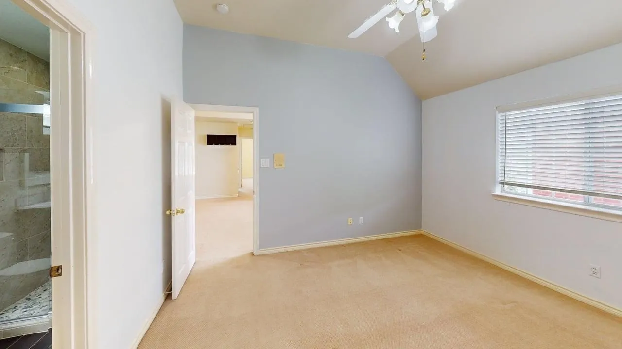 Vaulted ceiling with an attached ceiling fan, large window with blinds, carpeted flooring, and an adjoining bathroom with a glass-enclosed shower featuring tile walls and a built-in bench