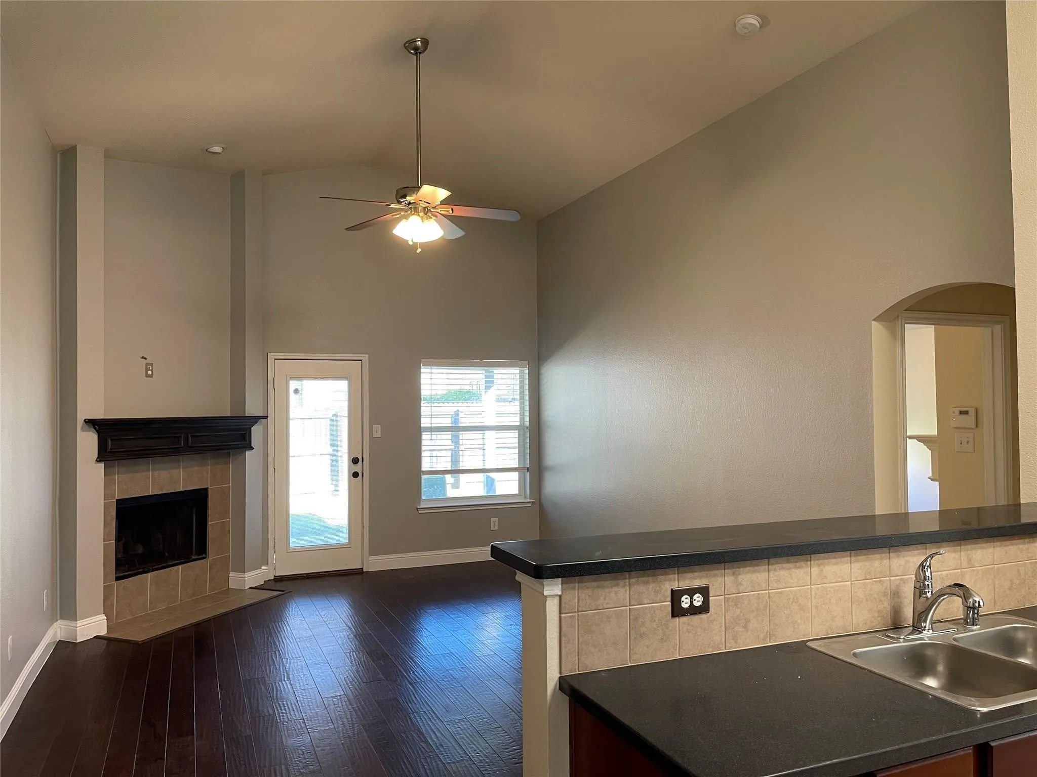 Vaulted ceiling, ceiling fan, fireplace with mantel and tile surround, wood-finish flooring, and kitchen passthrough with countertop