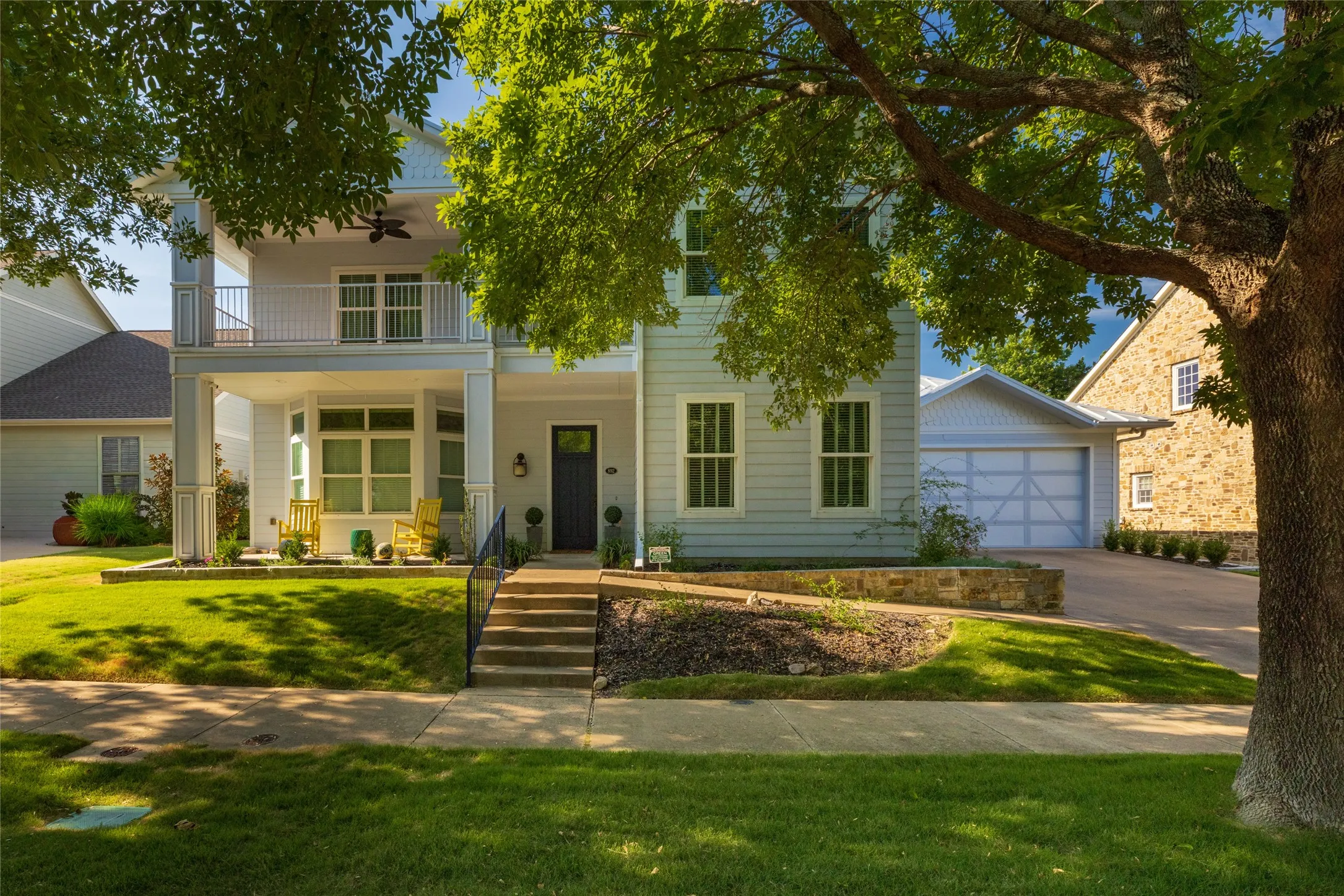 Light blue siding exterior with a covered porch, upper balcony, attached garage, and stone accent landscaping