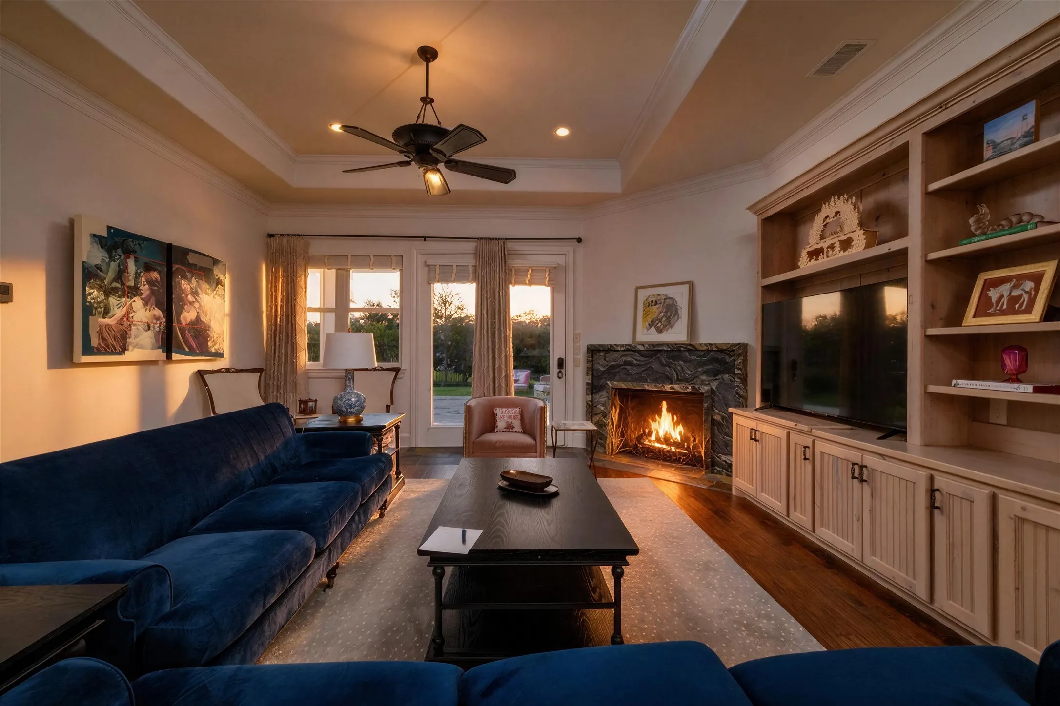 Tray ceiling with recessed lighting and a ceiling fan, stone-surround fireplace, built-in media center with shelving and lower cabinetry, wood-finish flooring, and French doors