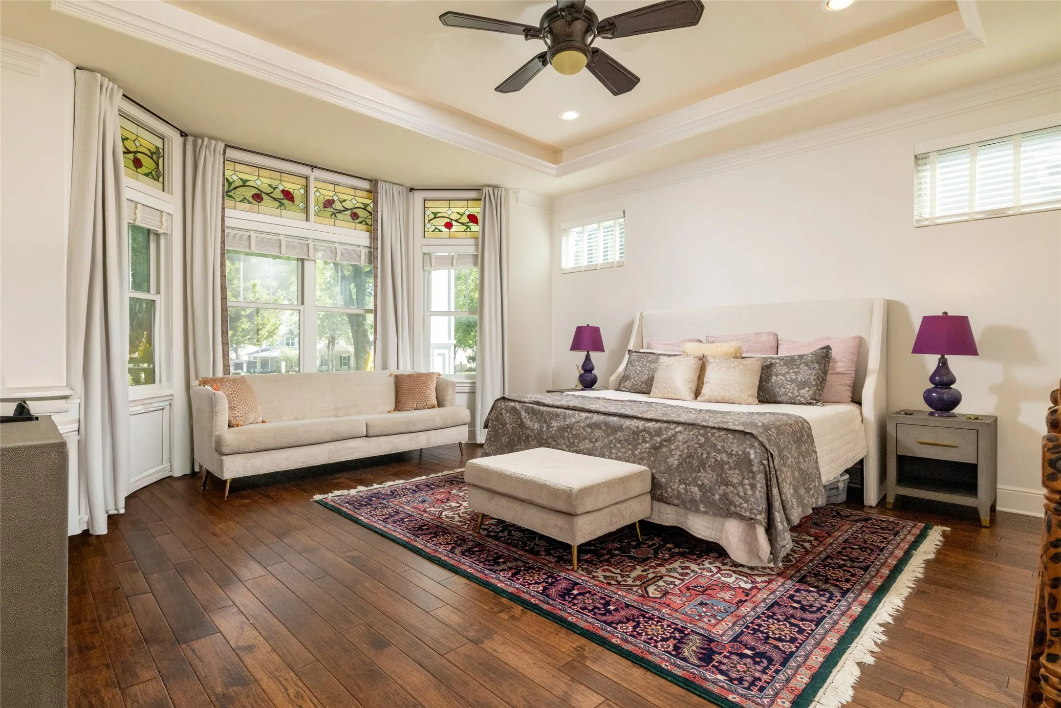 Hardwood flooring throughout, tray ceiling with recessed lighting and ceiling fan, bay window with decorative stained glass accents, and white window blinds