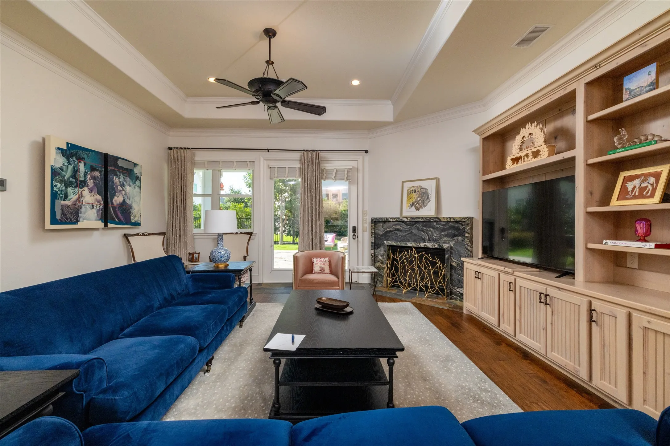 Tray ceiling with crown molding, dark wood-finish flooring, built-in entertainment center, stone fireplace surround with decorative screen, and ceiling fan with integrated lighting