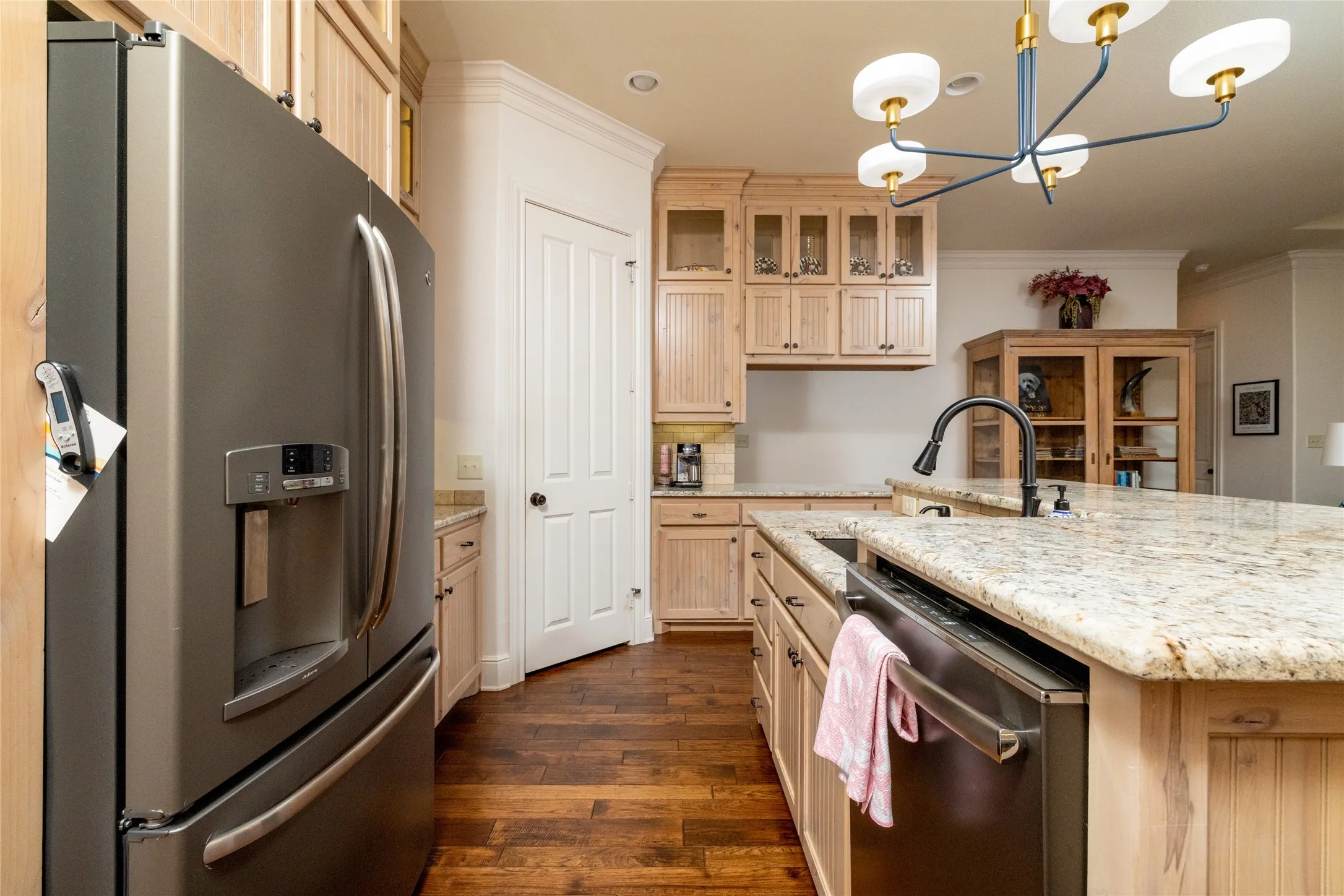 Spacious kitchen featuring stainless steel appliances, wood-finish flooring, light wood cabinetry with glass-front accents, and a large island with a composite stone countertop and integrated sink