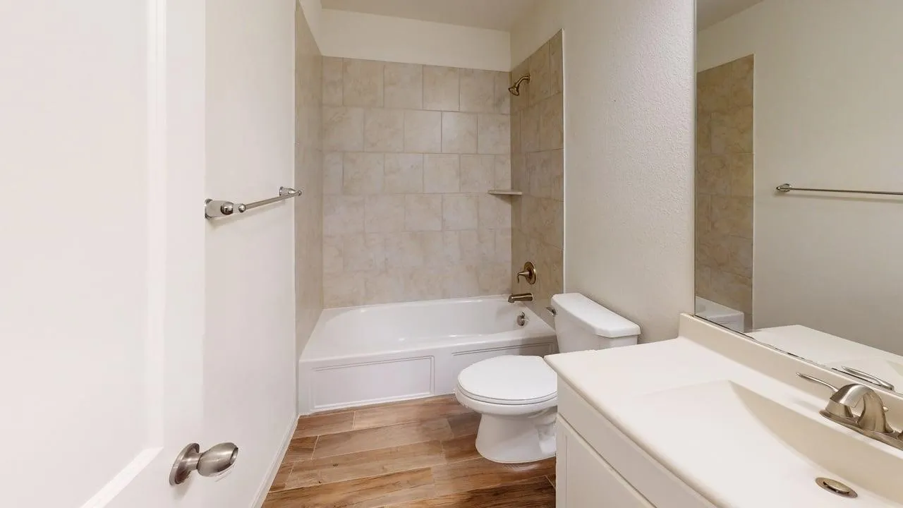 Bathroom with wood-finish flooring, a white vanity with an integrated sink, a large wall-mounted mirror, a white toilet, and a bathtub with tile surround