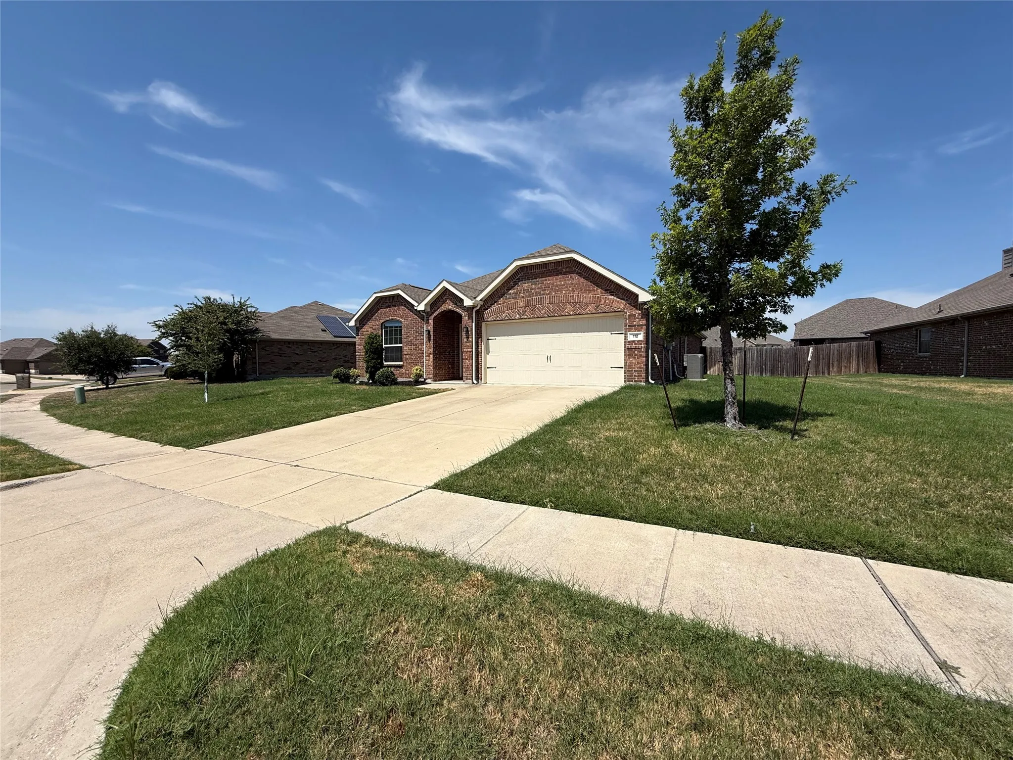Brick exterior featuring a two-car garage, mature trees, and a concrete driveway