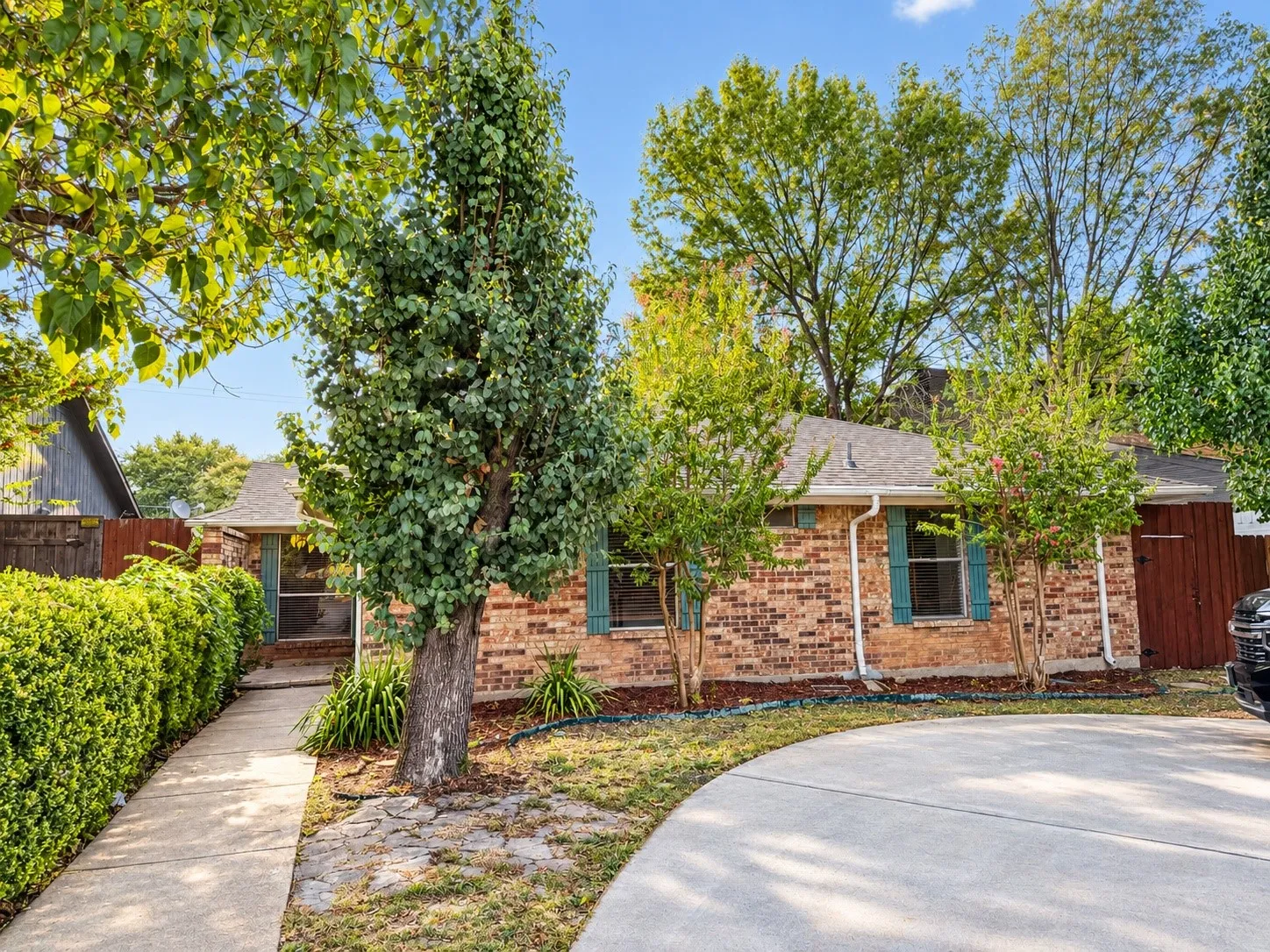 Brick exterior with contrasting green shutters, mature trees, manicured hedges, and a concrete driveway