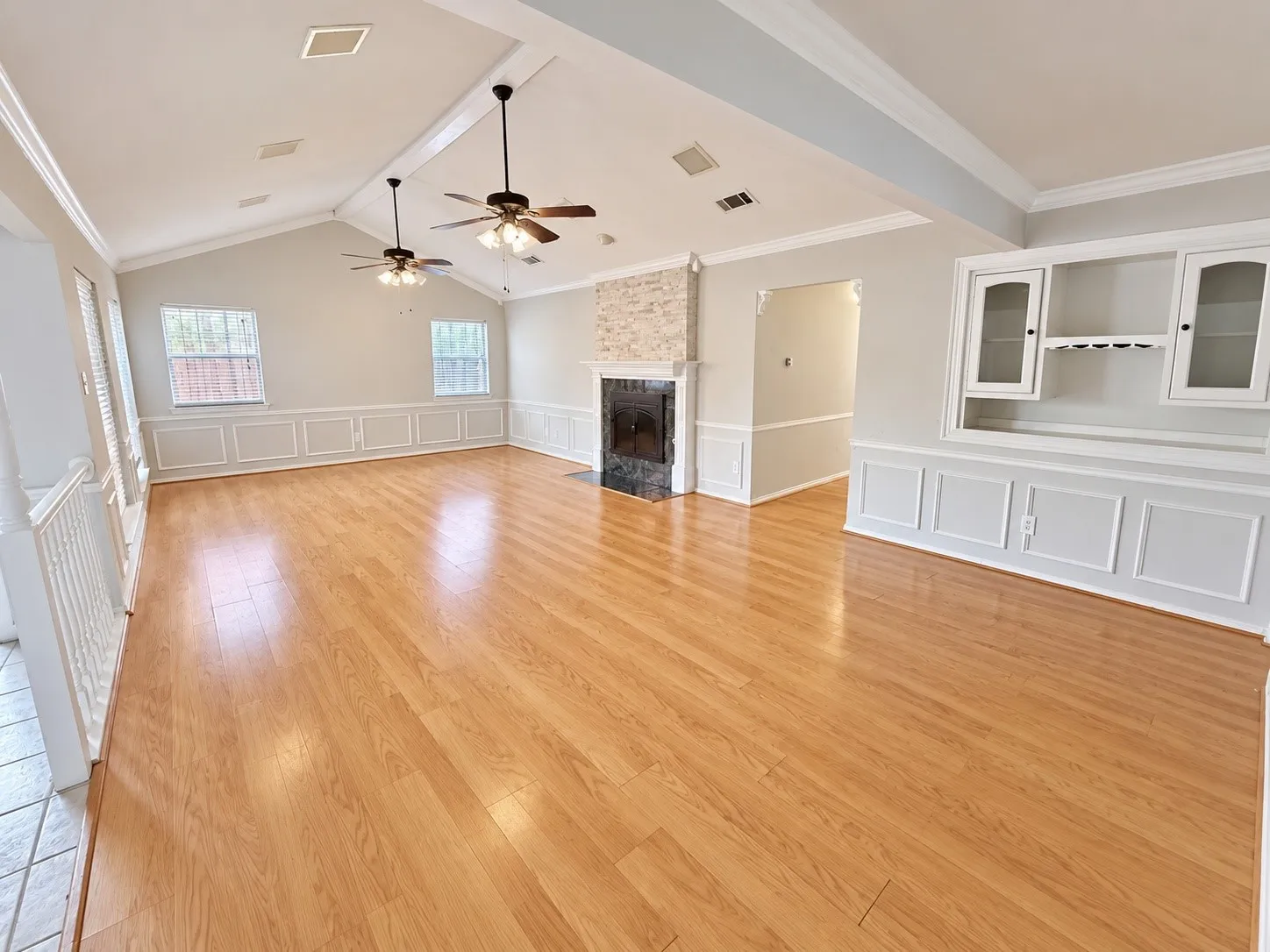 Vaulted ceiling with exposed beam, wood-finish flooring, built-in cabinetry with glass-front doors, stone-clad fireplace with mantel, and decorative wainscoting