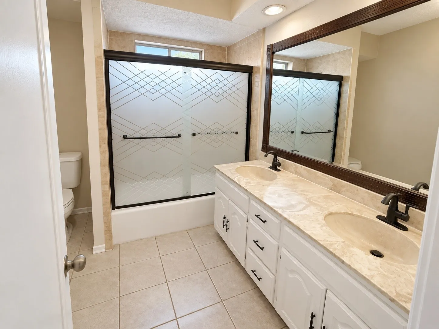 Dual vanity with integrated sinks, framed mirror, tiled flooring, and a shower-tub combination with patterned glass doors