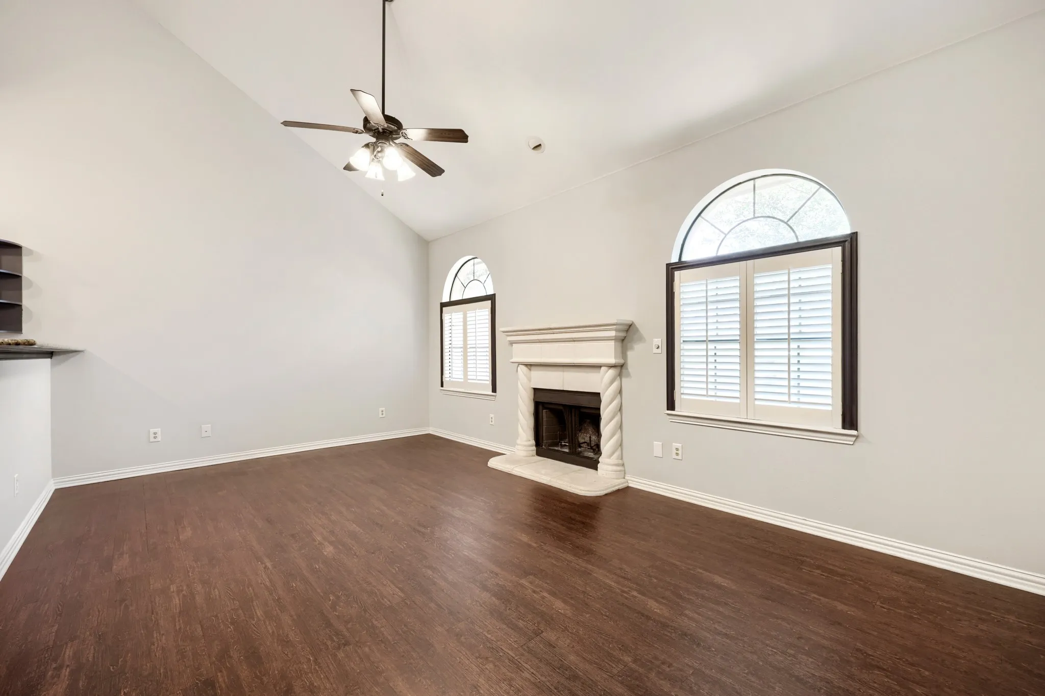 Vaulted ceiling with a ceiling fan, fireplace with a decorative mantel, arched windows with plantation shutters, and wood-finish flooring