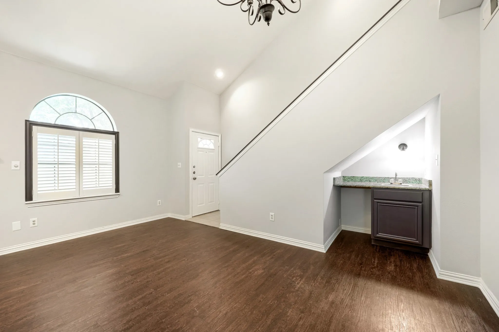 Wood-finish flooring throughout, arched window with plantation shutters, front entry door with transom window, built-in wet bar with cabinet and sink, and a staircase with an ornamental chandelier