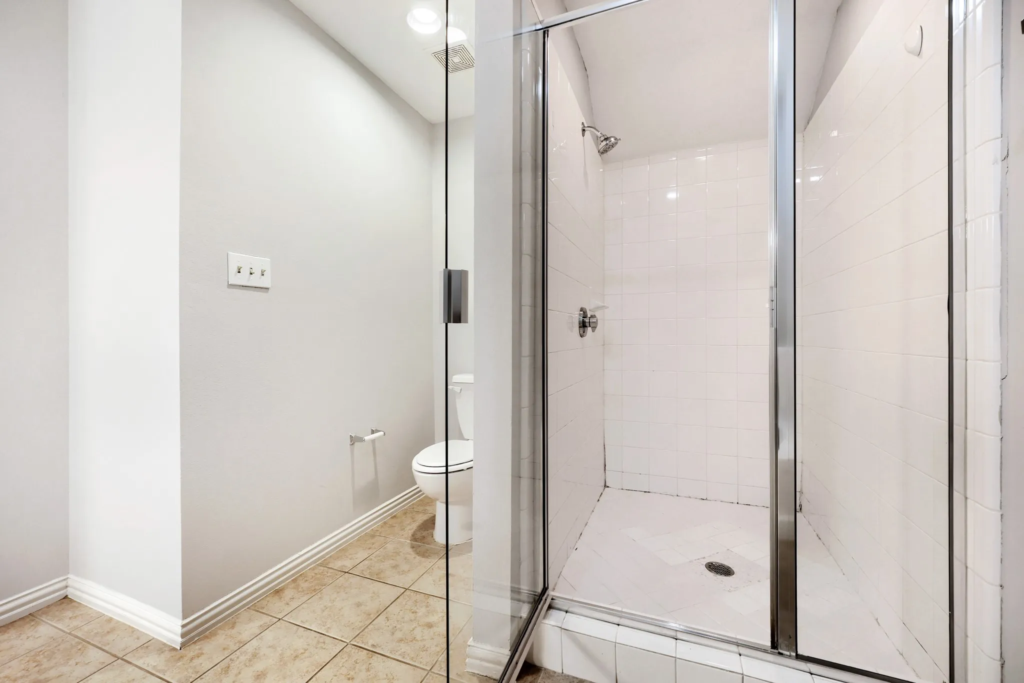 Glass-enclosed shower with white tiled walls and floor, complemented by a toilet and tiled flooring