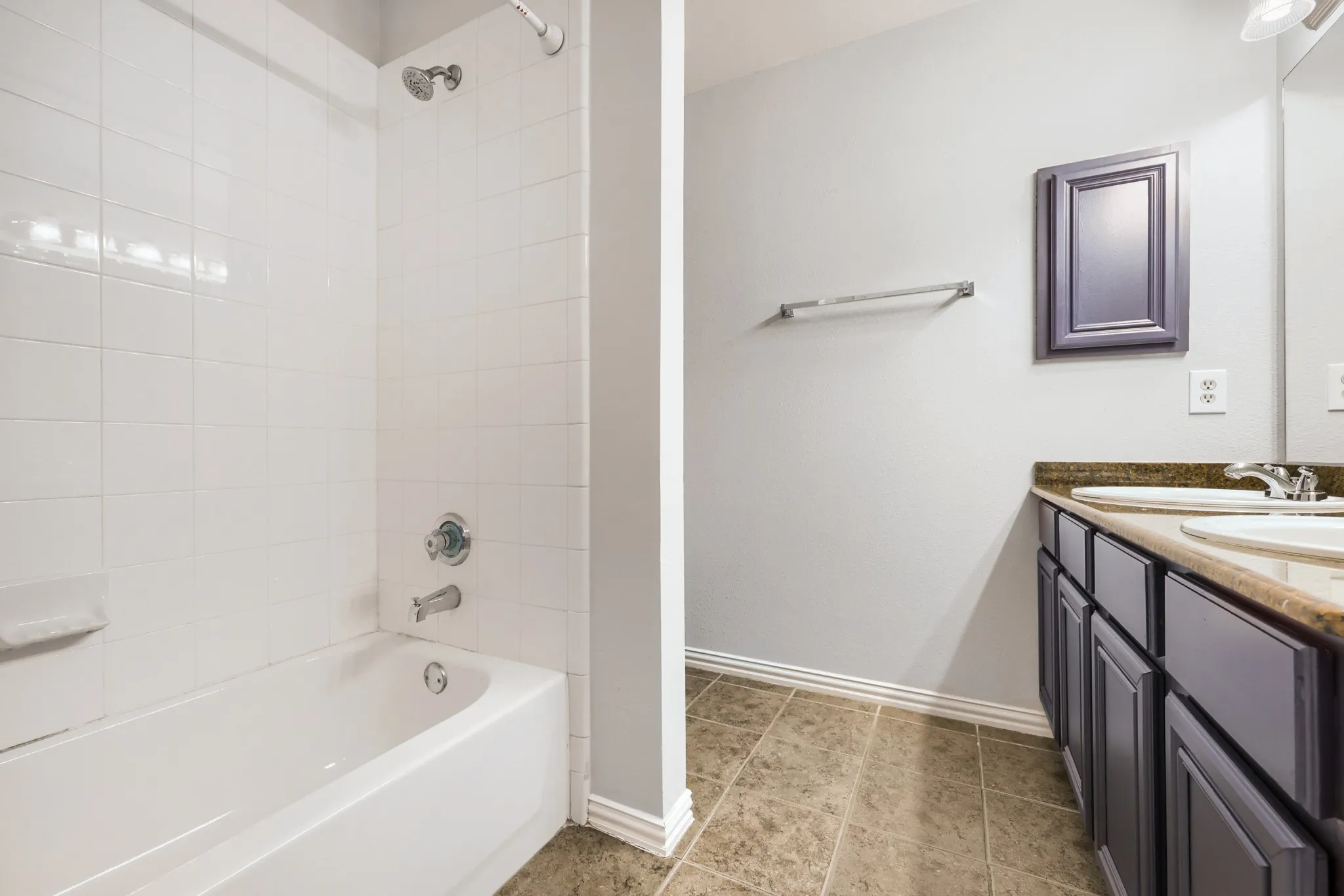 Bathroom featuring a white bathtub with tile surround, dual sink vanity with stone-look countertop, dark cabinetry, and tile flooring