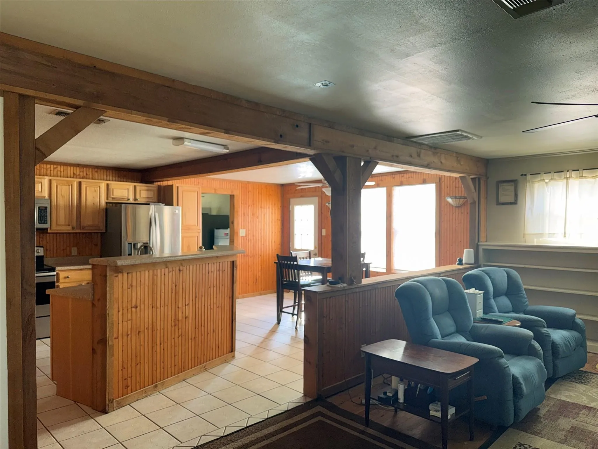 Open concept common area featuring wood-beam accents, a kitchen with wood cabinetry and stainless steel appliances, a breakfast bar, and tile flooring
