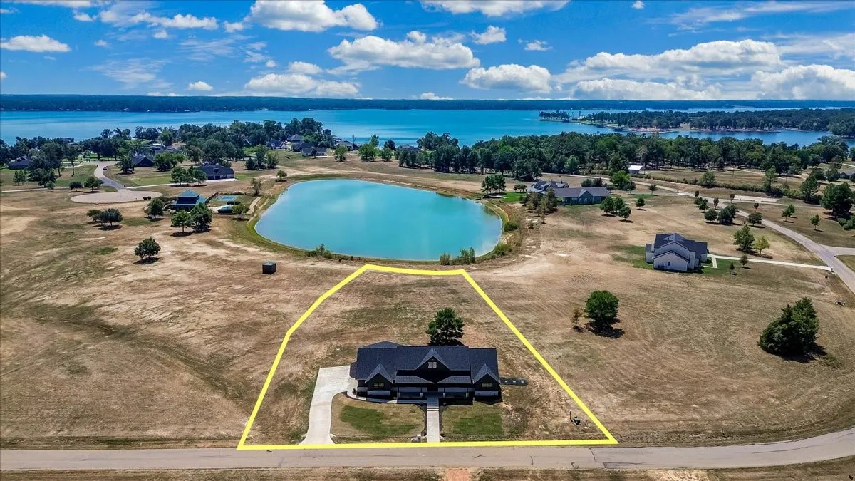 Expansive property featuring a modern residence with a dark roof and light-colored driveway, a nearby pond, and distant lake scenery