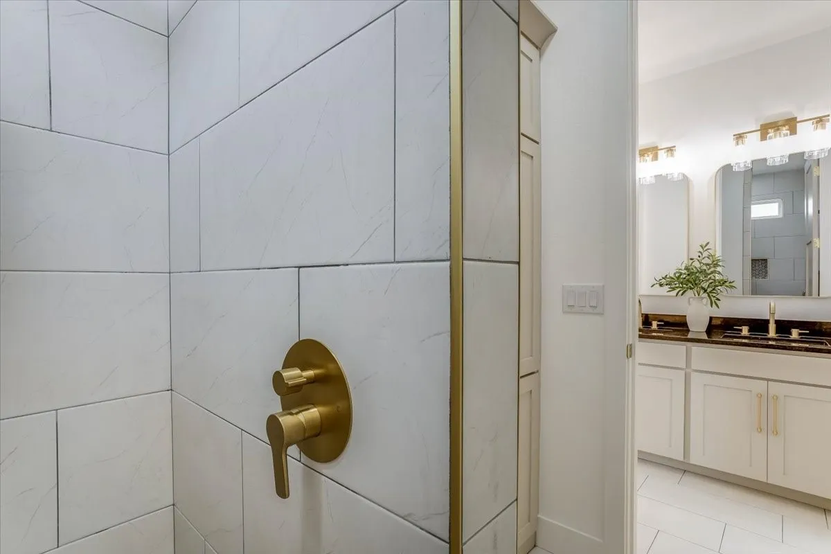 White tiled shower with brass fixtures, built-in storage cabinet, dual vanity with dark countertops, and gold-tone sconce lighting