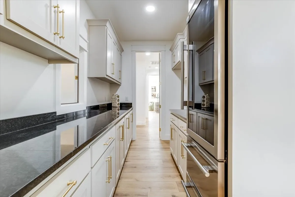 Galley kitchen featuring extensive dark countertops, light-toned cabinetry with gold-finish hardware, integrated stainless steel appliances, and wood-finish flooring