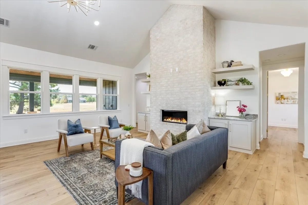 Vaulted ceiling with a contemporary chandelier, wood-finish flooring, brick-faced fireplace with an electric insert, built-in cabinetry with floating shelves, and multiple large windows