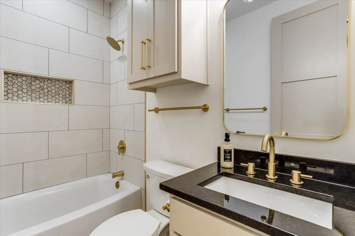 White subway tile shower surround with a decorative niche, a built-in bathtub, a vanity with a dark countertop, an undermount sink, and a gold-framed mirror