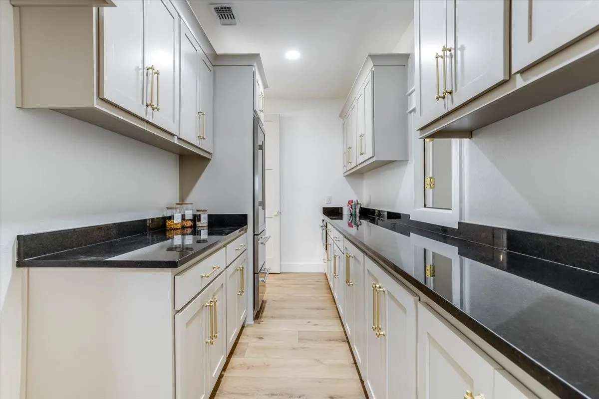 Galley-style kitchen with light wood-finish flooring, white cabinetry, dark countertops, and gold-tone hardware