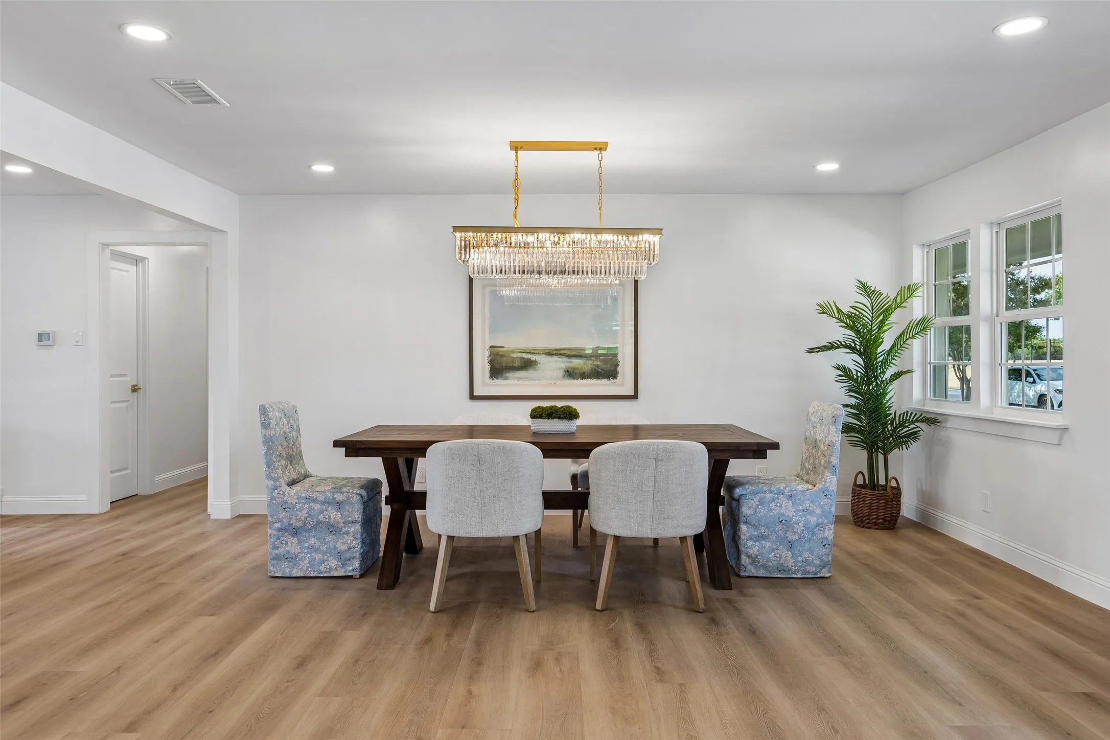The dining area sits within the great room, anchored by a striking linear chandelier and framed by natural light from the windows and glass paneled front doors. Engineered wood flooring continues seamlessly from the adjoining spaces, creating an easy flow for both everyday meals and entertaining