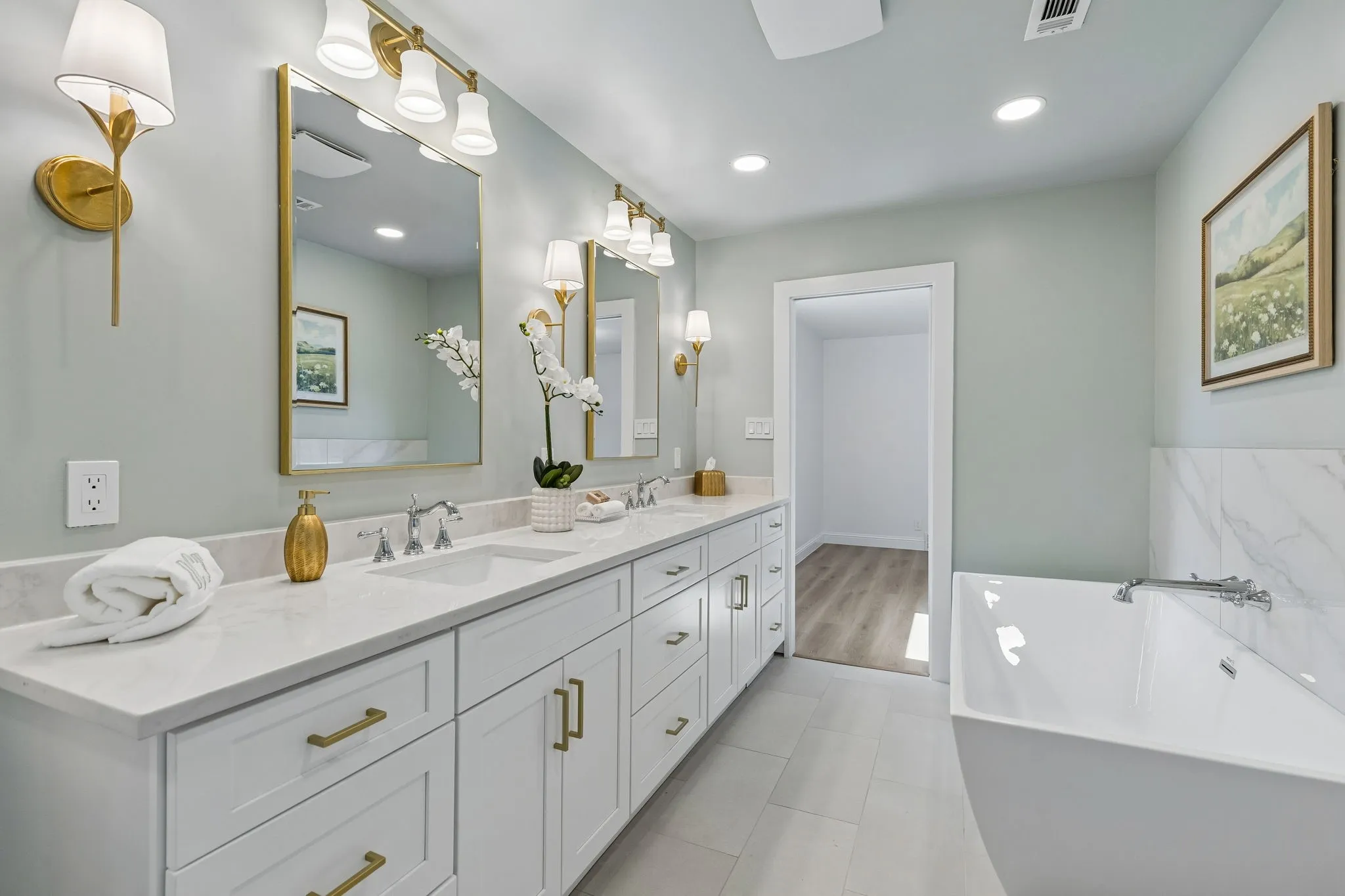 Luxurious primary bath with dual vanities, brass framed mirrors, and elegant sconce lighting, plus a walk in closet just beyond.