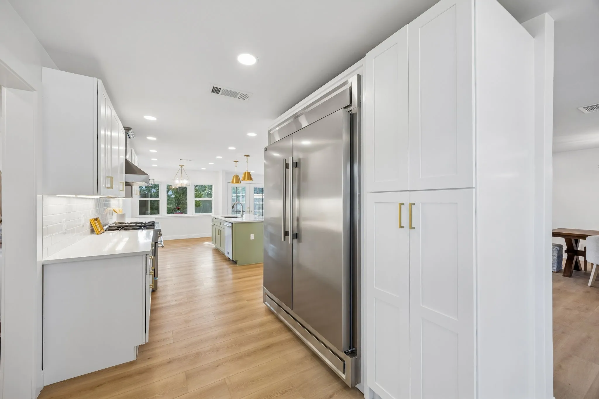 Chef's kitchen with a full size stainless refrigerator, crisp white cabinetry with gold pulls, and an open flow into the sunlit living spaces beyond.