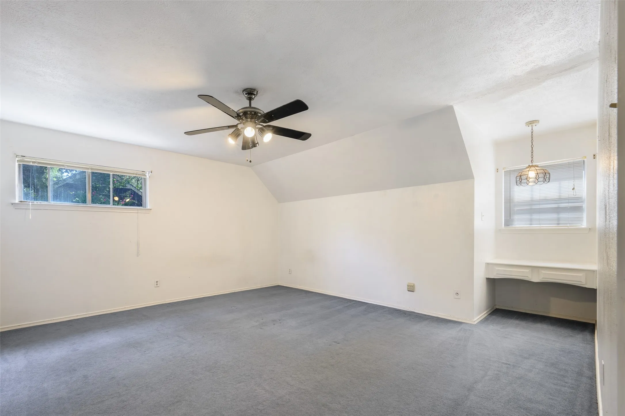 Expansive room featuring sloped ceilings, a ceiling fan with integrated lighting, a built-in desk, and a decorative pendant light fixture