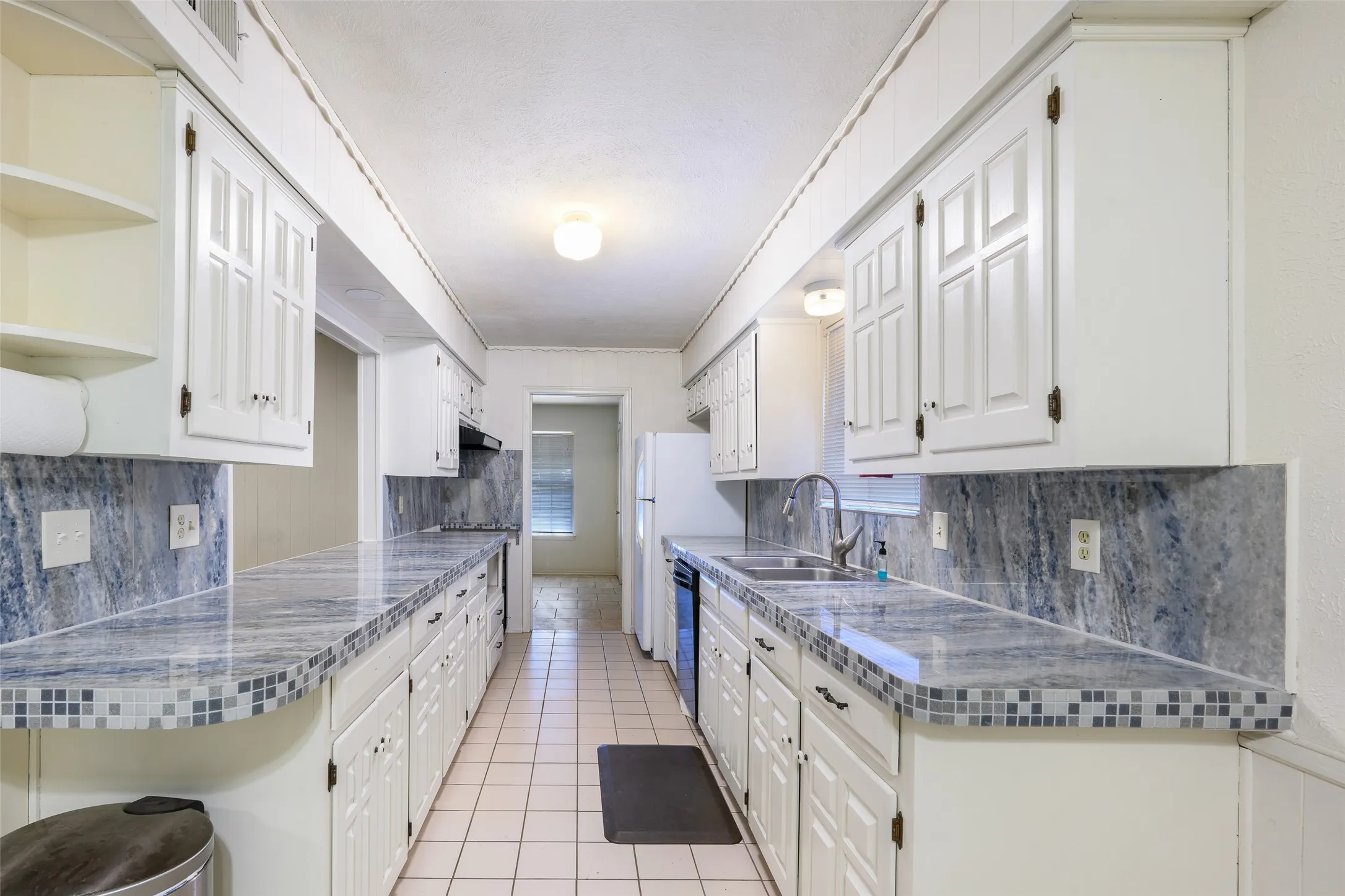 White cabinetry with dark hardware, stone-look countertops with tile trim, ceramic tile flooring, and integrated dual basin sink