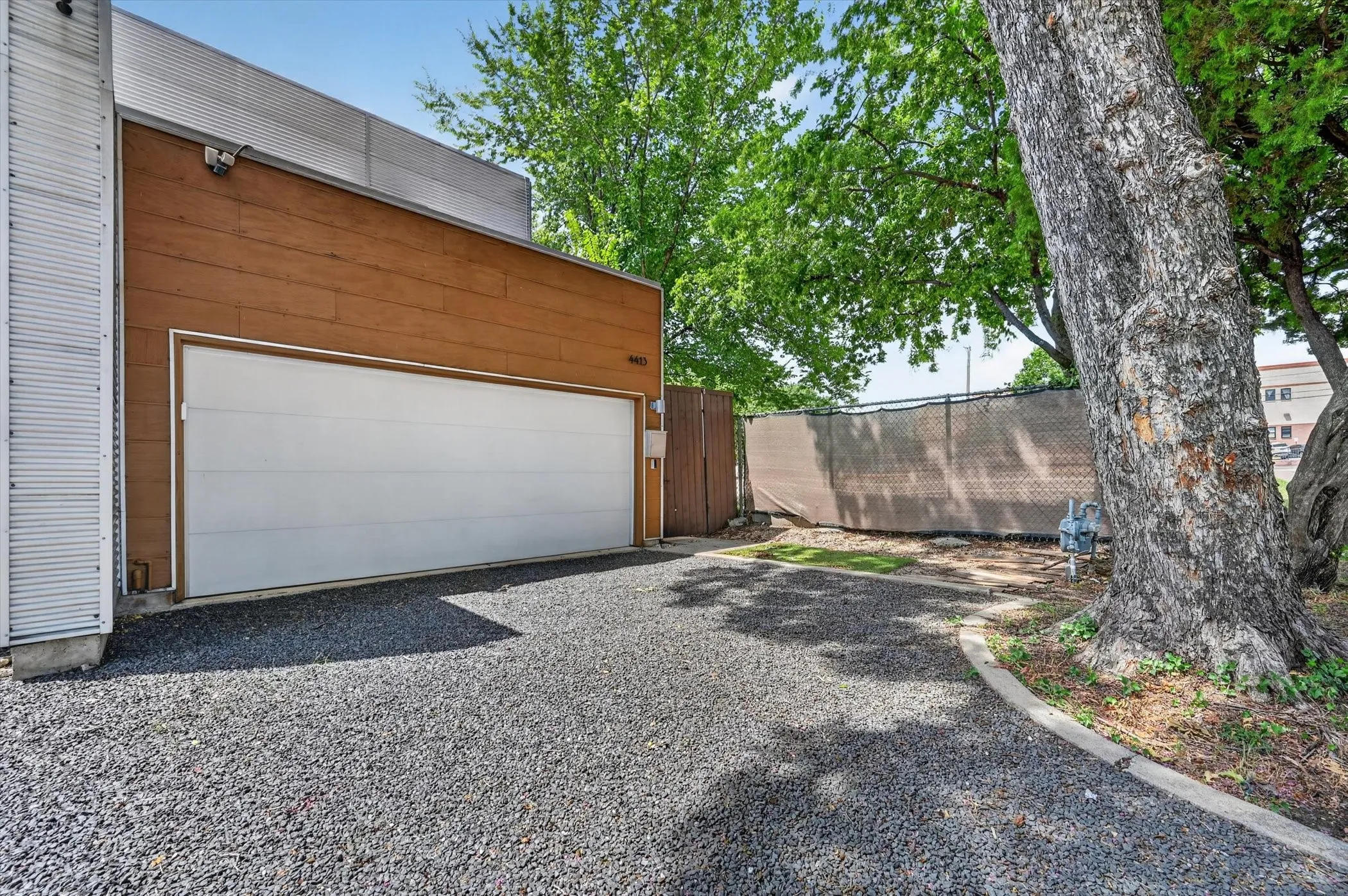 Wood-paneled facade with a wide garage door, gravel driveway, and mature trees
