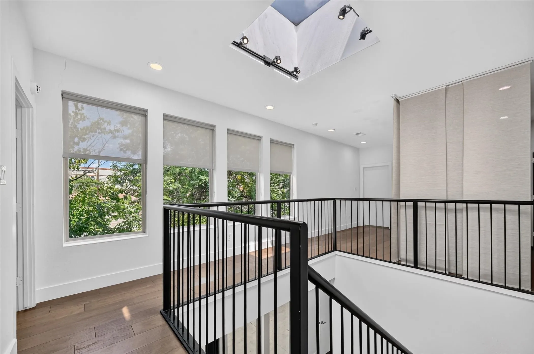 Upper level landing featuring wood-finish flooring, a black metal railing, multiple windows with roller shades, recessed lighting, and a skylight with track lighting