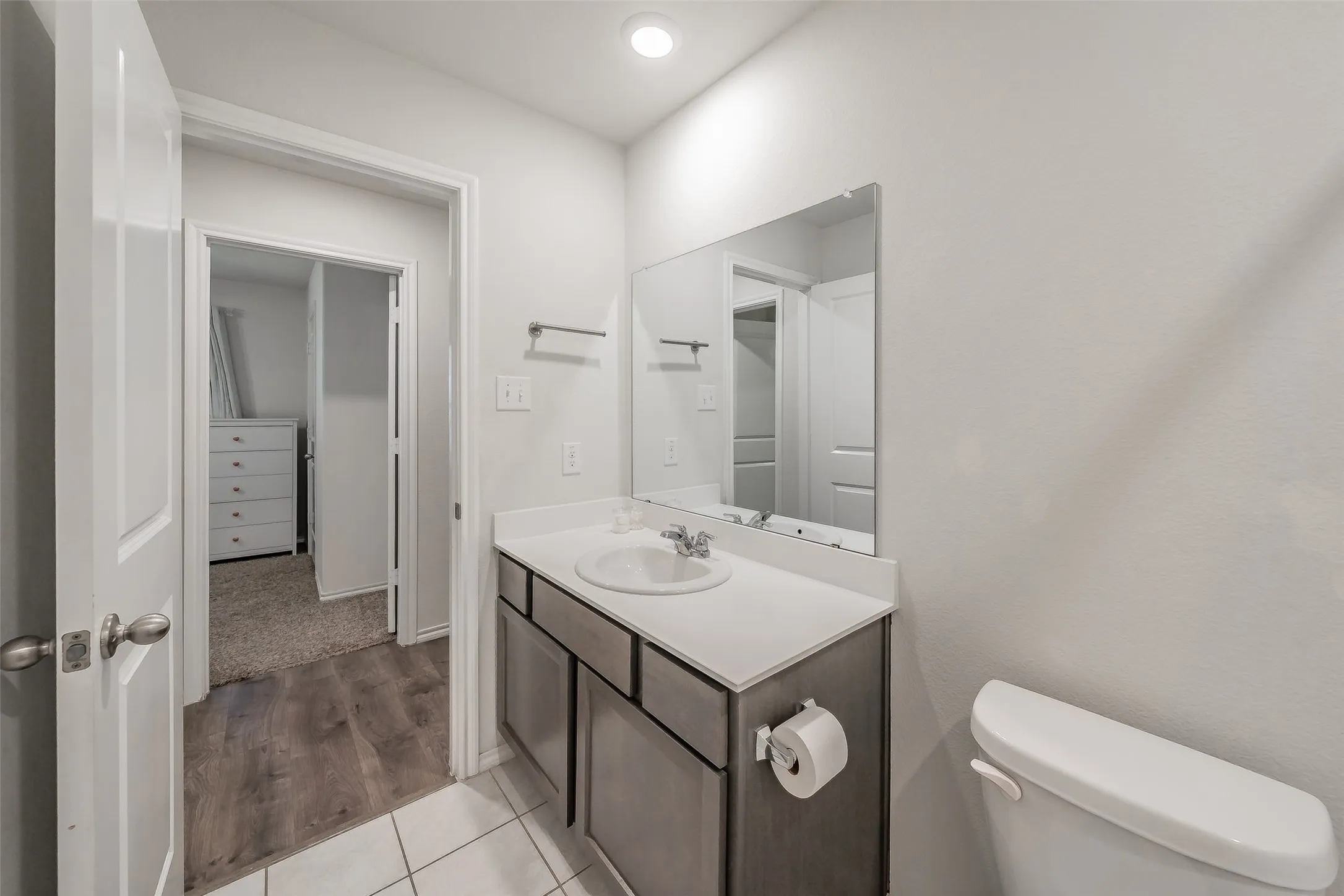 Bathroom featuring a single vanity with a white countertop, chrome faucet, wall-mounted mirror, and white tile flooring