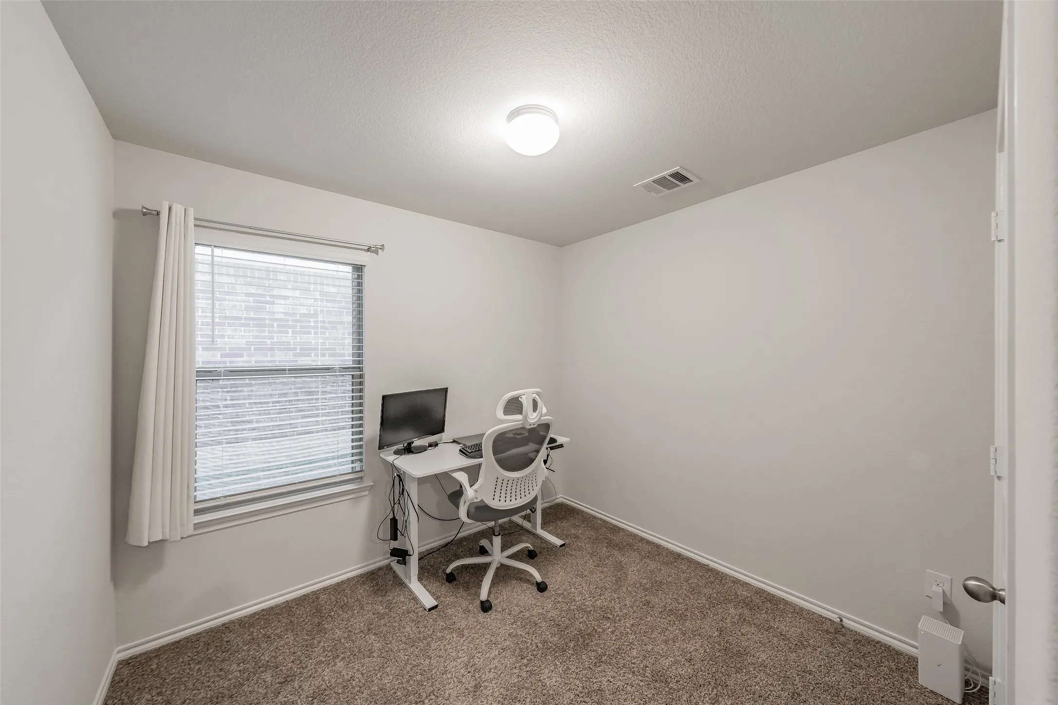 Carpeted bedroom being used as an office with a single window, overhead lighting, and a white door