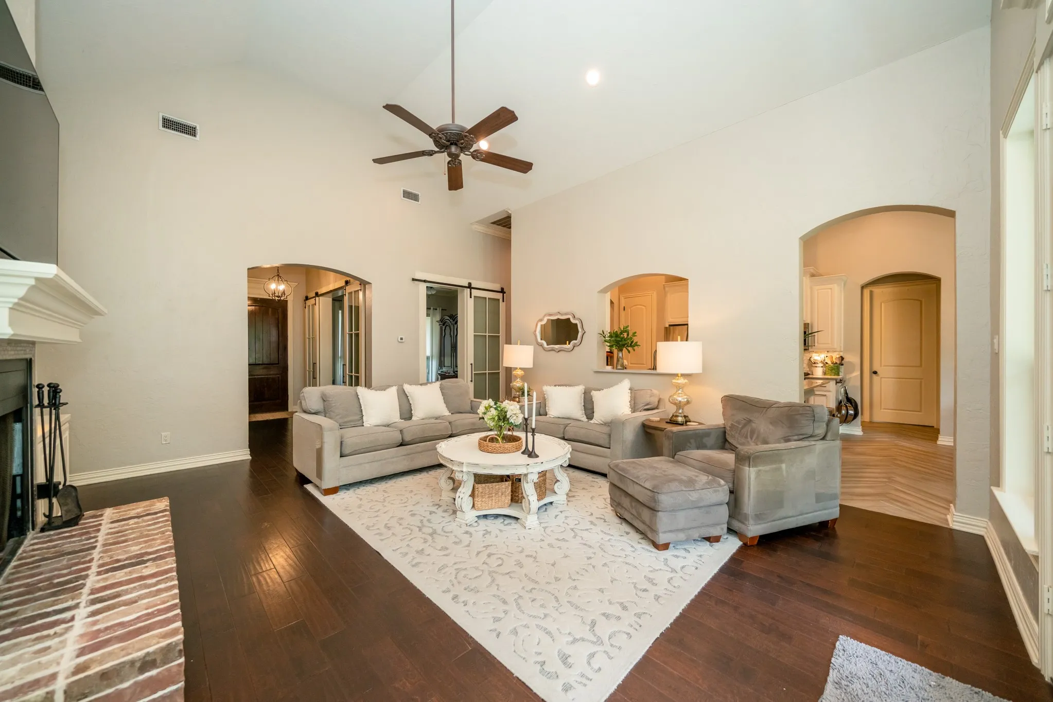 Vaulted ceiling with a ceiling fan, dark wood-finish flooring, fireplace with a brick hearth, arched doorways, and sliding barn doors