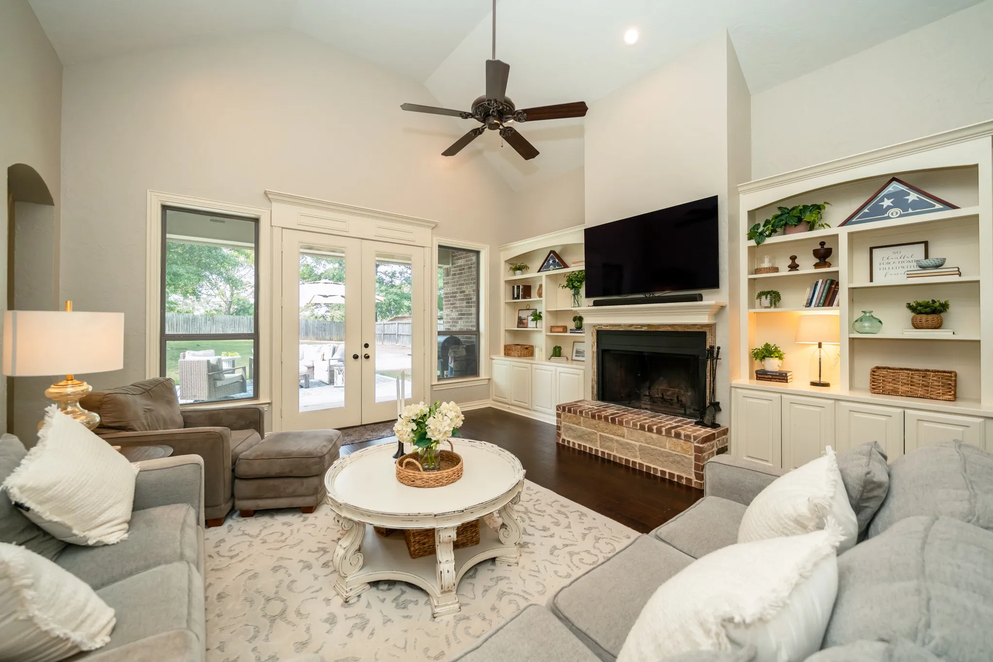 Vaulted ceiling with a ceiling fan, built-in shelving, brick fireplace with a mantel, dark wood-finish flooring, and French doors leading to an outdoor space