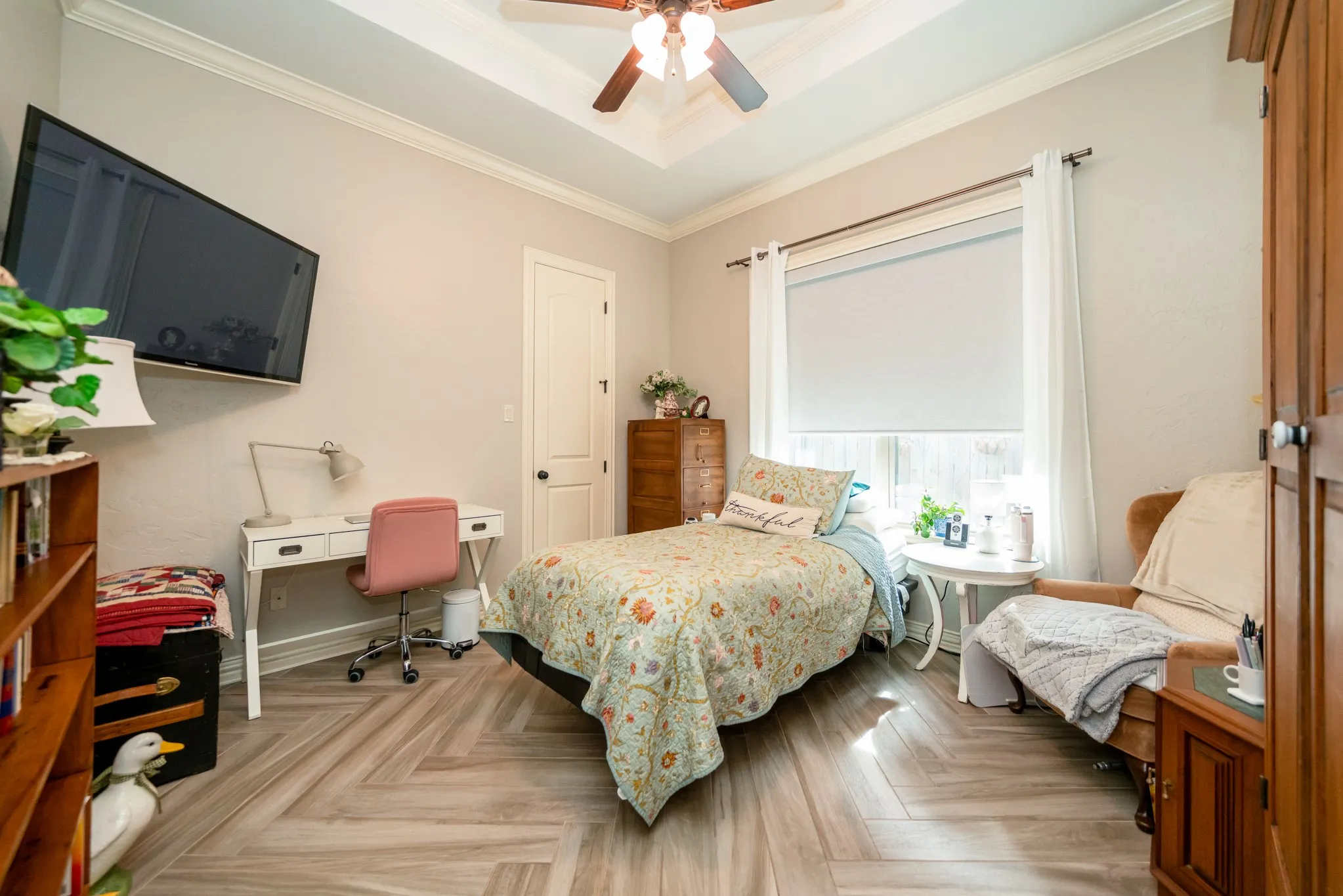 Tray ceiling with a ceiling fan, crown molding, wood-finish flooring in a herringbone pattern, and a window with roller shades