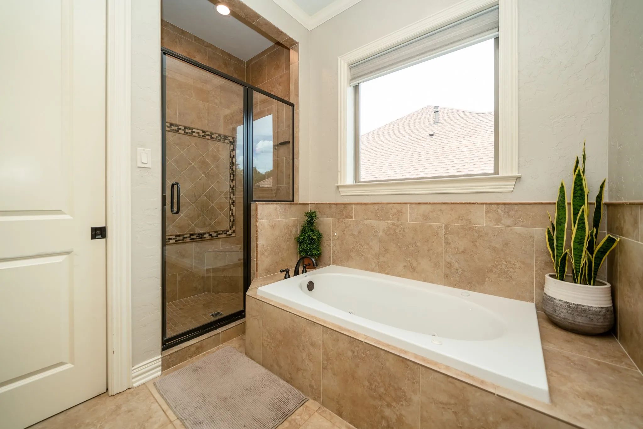 Built-in soaking tub with tile surround, walk-in shower with glass enclosure, tiled walls, and a window with a roller shade