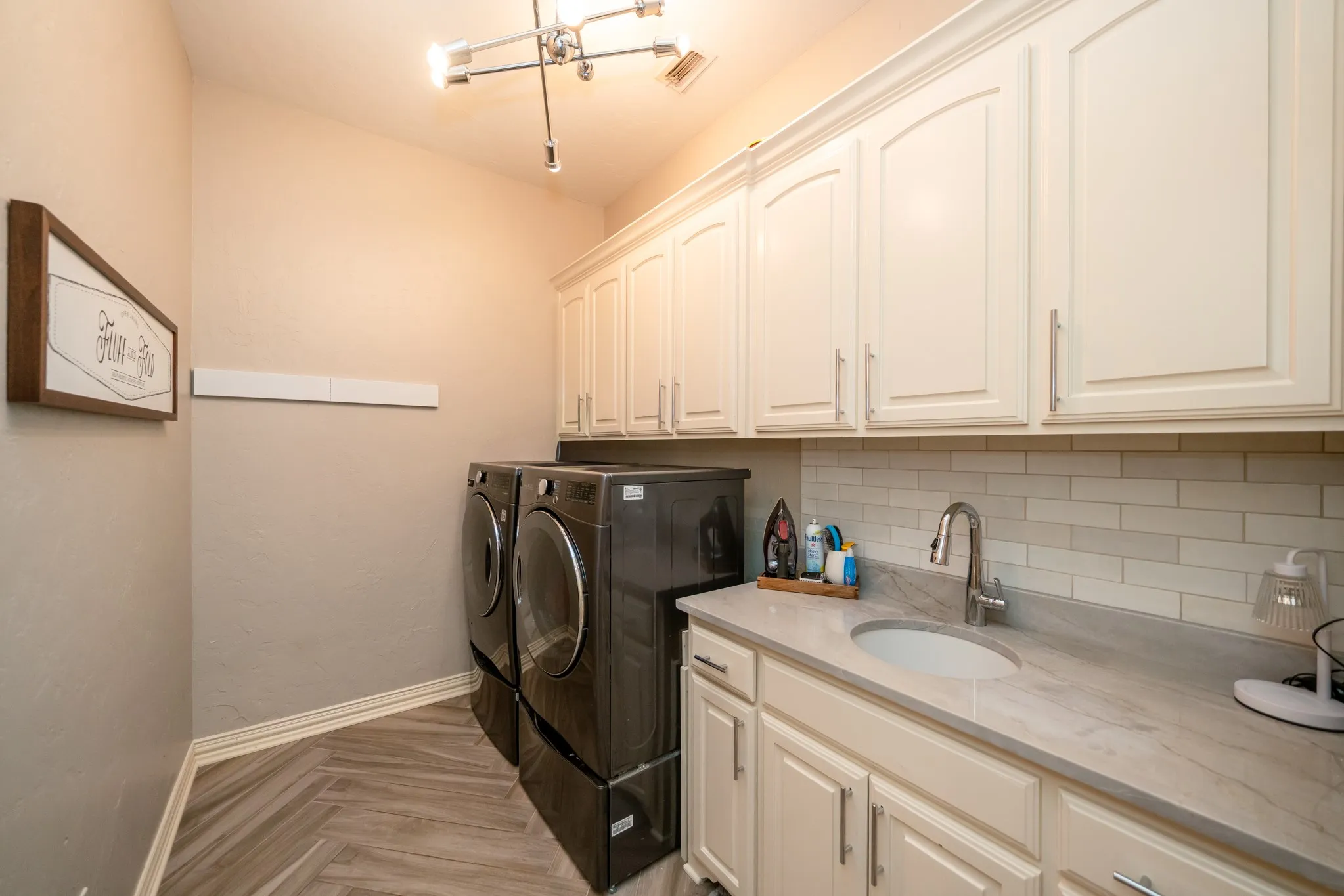 Herringbone pattern wood-finish flooring, white shaker-style cabinetry with chrome hardware, subway tile backsplash, built-in sink with a gooseneck faucet, and track lighting