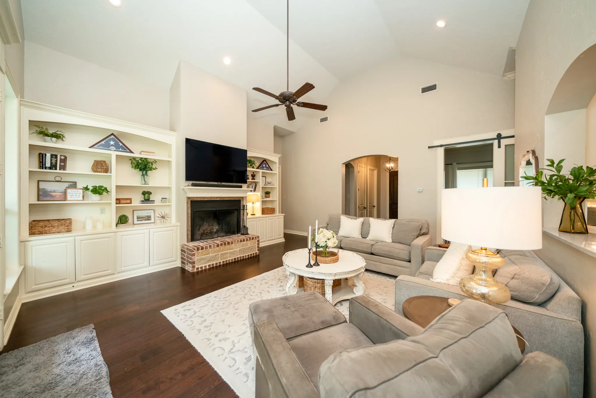 Vaulted ceiling with recessed lighting, wood-finish flooring, built-in shelving, brick fireplace surround, and a barn door entry