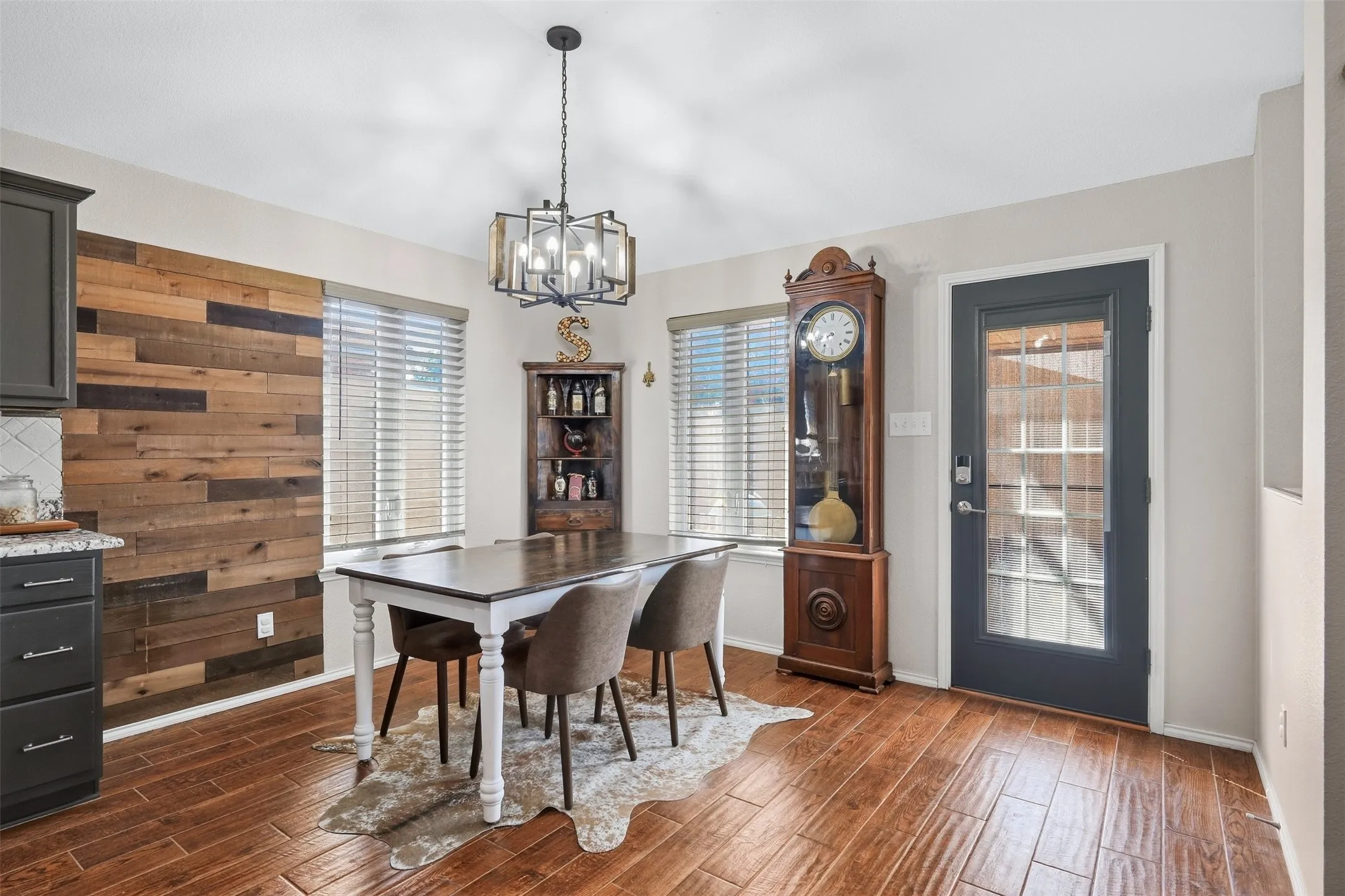 Wood-finish flooring, accent wall with varied wood paneling, contemporary chandelier, multiple windows with blinds, and a glass-paneled exterior door