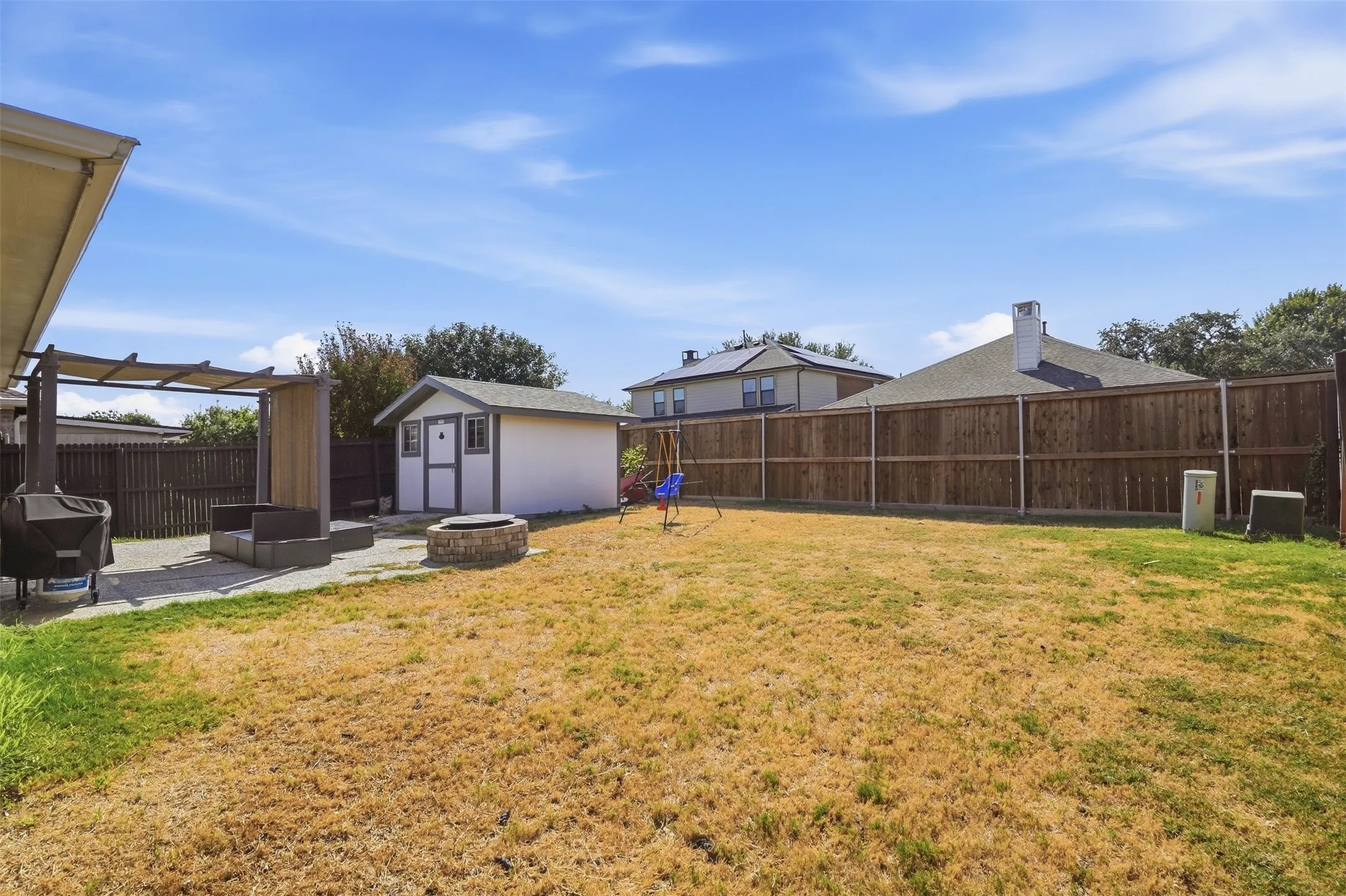 Expansive backyard featuring a privacy fence, concrete patio, pergola, storage shed, and a stone-finish fire pit