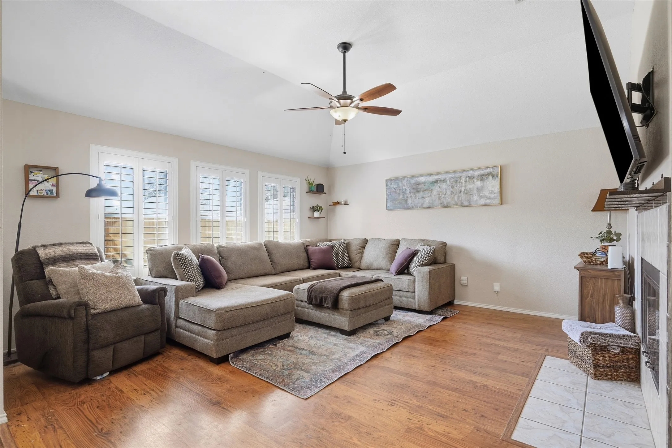 Vaulted ceiling with a ceiling fan, wood-finish flooring, multiple windows with plantation shutters, built-in wall shelves, and a tile fireplace surround